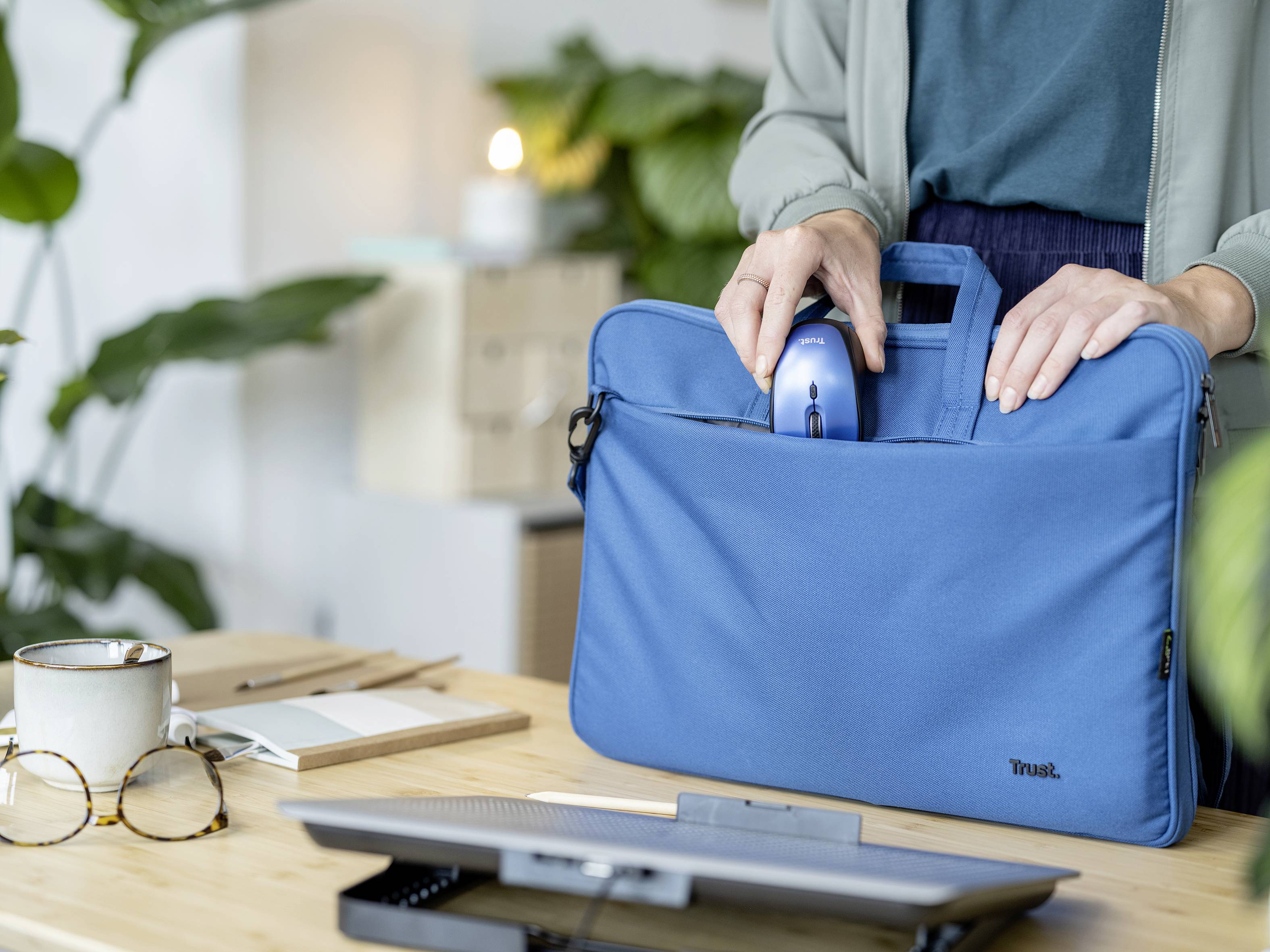 Person steckt eine Computermaus in eine blaue Laptoptasche auf einem Tisch. Im Hintergrund sind Pflanzen und Kartons zu sehen.