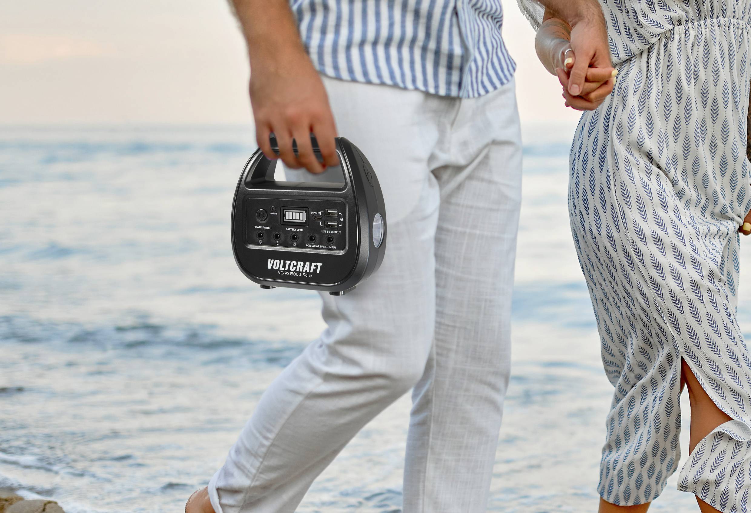 Ein Mann und eine Frau spazieren am Strand entlang. Der Mann hält ein tragbares Gerät der Marke 'Voltcraft' in der Hand.