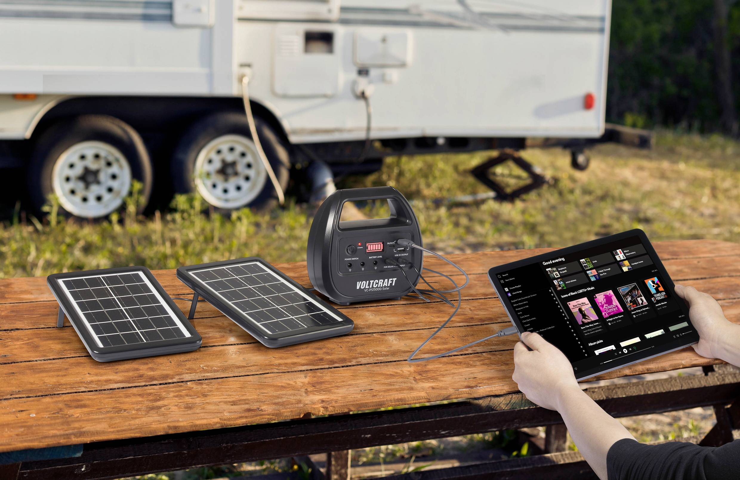Solarpanels und Ladegerät auf einem Holztisch, verbunden mit einem Tablet, im Hintergrund ein Wohnwagen. Mobil laden unterwegs.