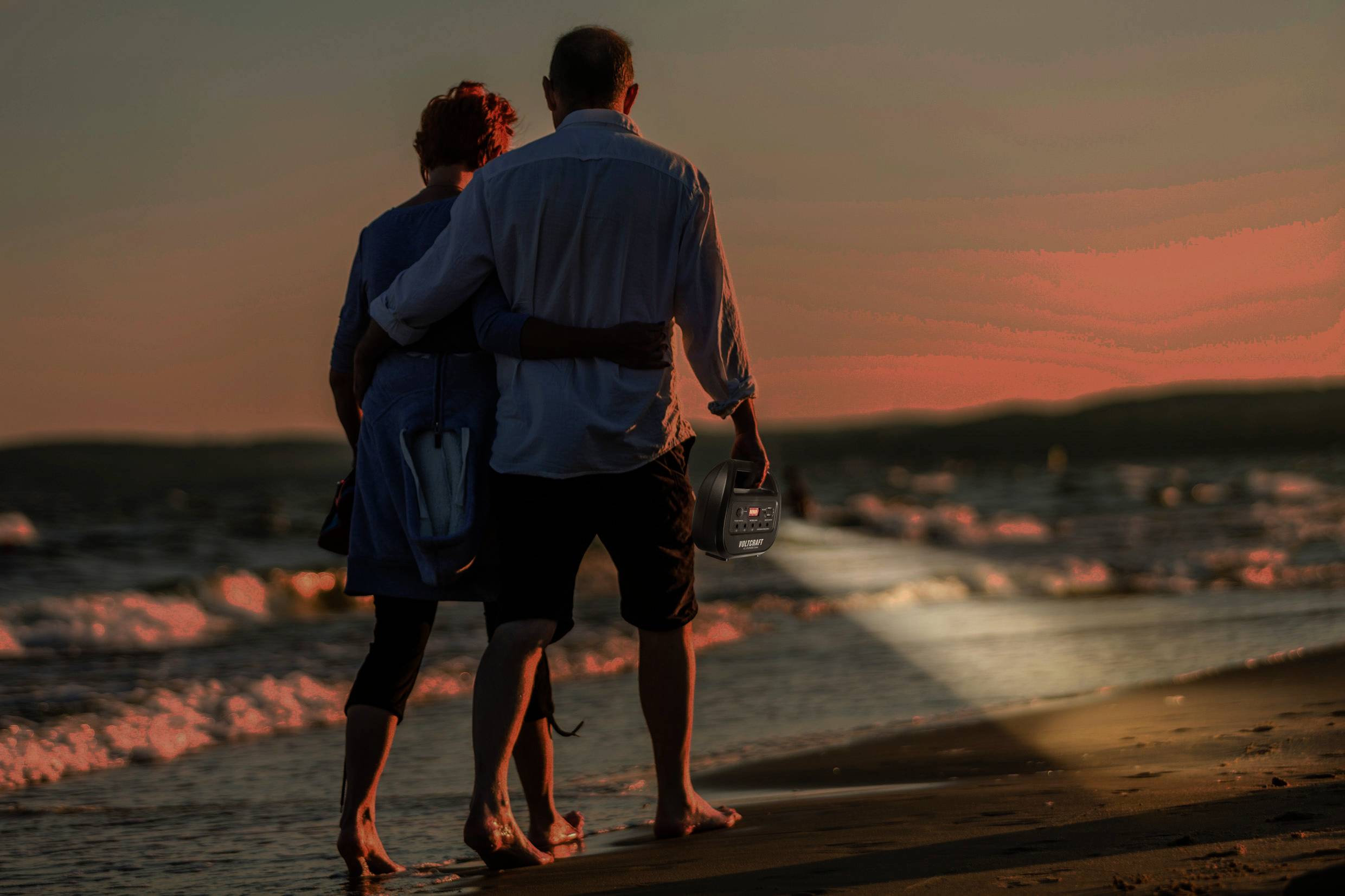 Ein älteres Paar spaziert bei Sonnenuntergang am Strand entlang. Der Mann hält eine kleine leuchtende Laterne in der Hand.