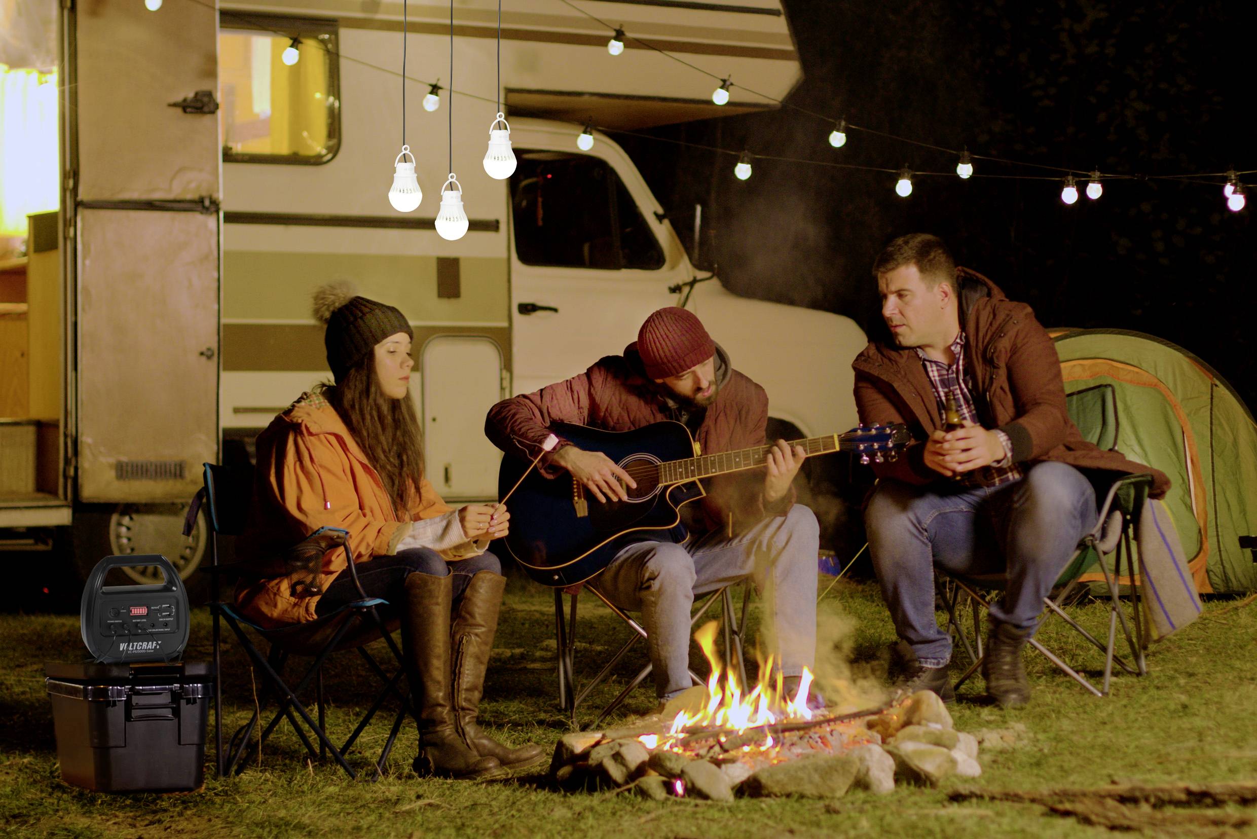 Drei Personen sitzen um ein Lagerfeuer bei einem Wohnmobil. Eine Person spielt Gitarre, die anderen hören zu. Im Hintergrund sind Lichterketten.