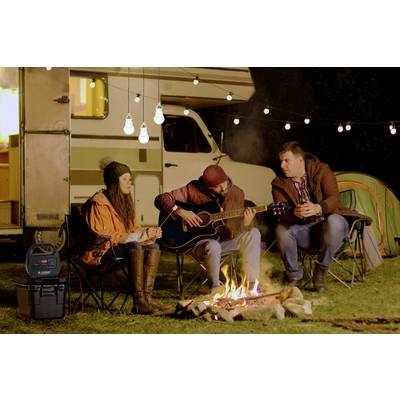 Drei Personen sitzen um ein Lagerfeuer bei einem Wohnmobil. Eine Person spielt Gitarre, die anderen hören zu. Im Hintergrund sind Lichterketten.