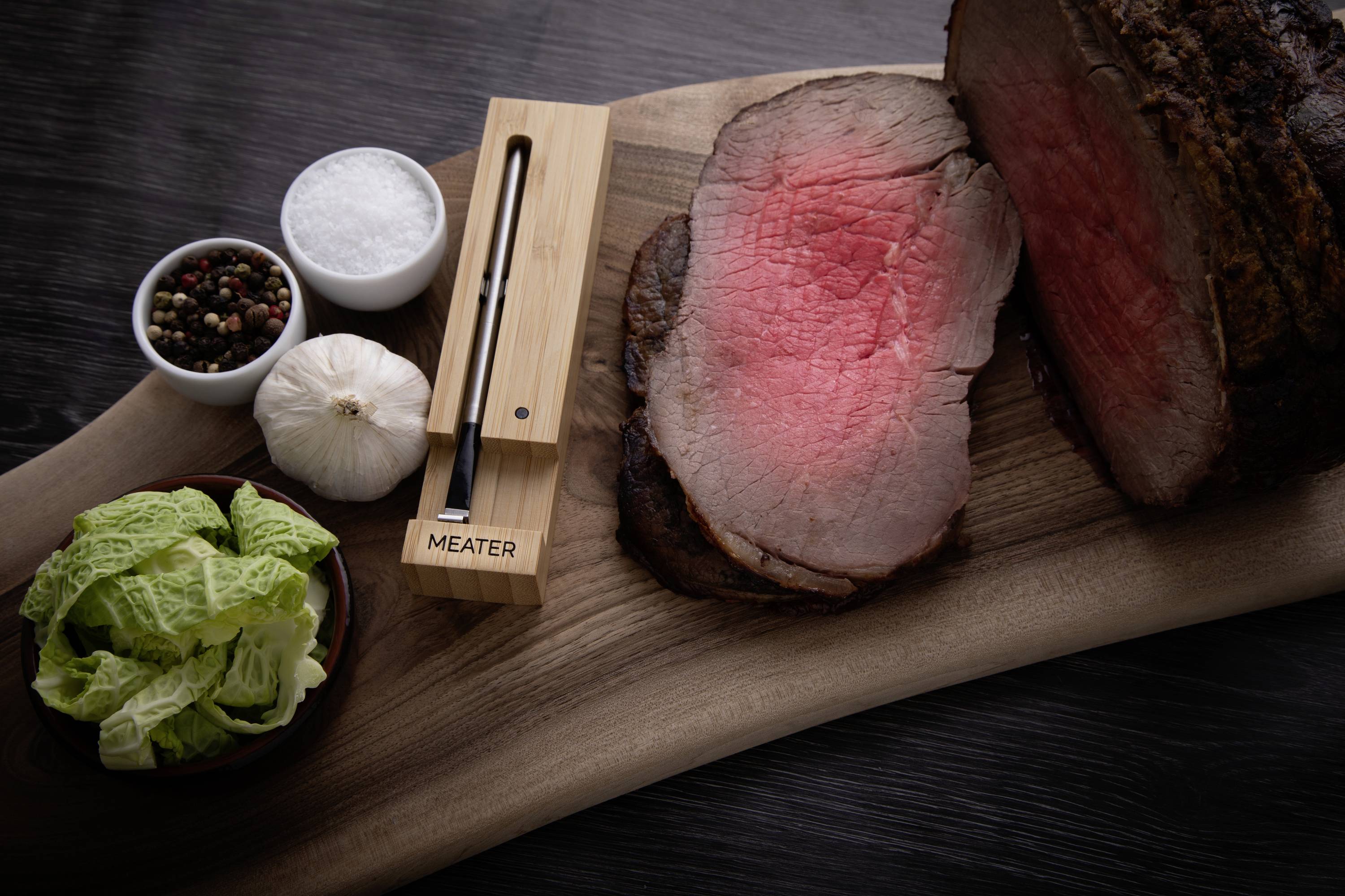 'Slice eines medium gebratenen Roastbeefs auf Holzbrett, mit Salz, Pfeffer, Knoblauch, Salat und Fleischthermometer als Dekoration.'