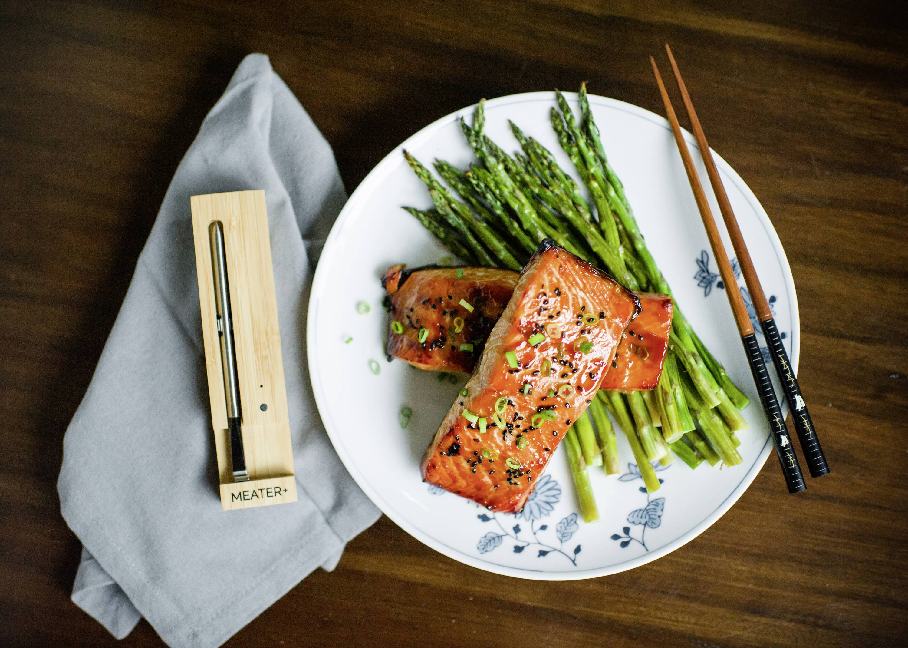 Gericht mit gegrilltem Lachs und Spargel auf einem weißen Teller, daneben ein Fleischthermometer. Essstäbchen liegen rechts auf dem Tisch.