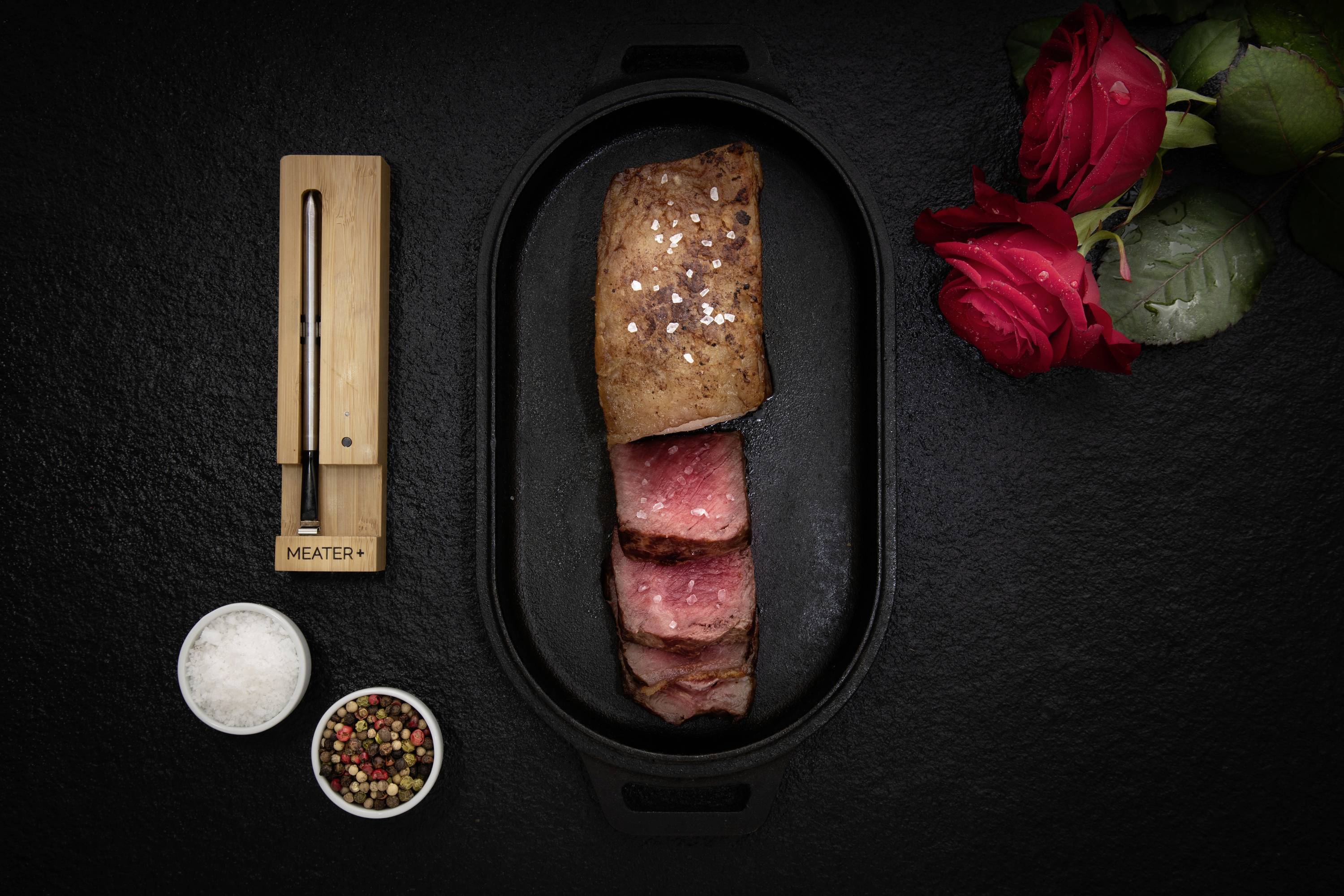 Ein Medium-Steak, halb aufgeschnitten, in einer schwarzen Pfanne. Daneben ein digitaler Fleischthermometer und zwei rote Rosen.