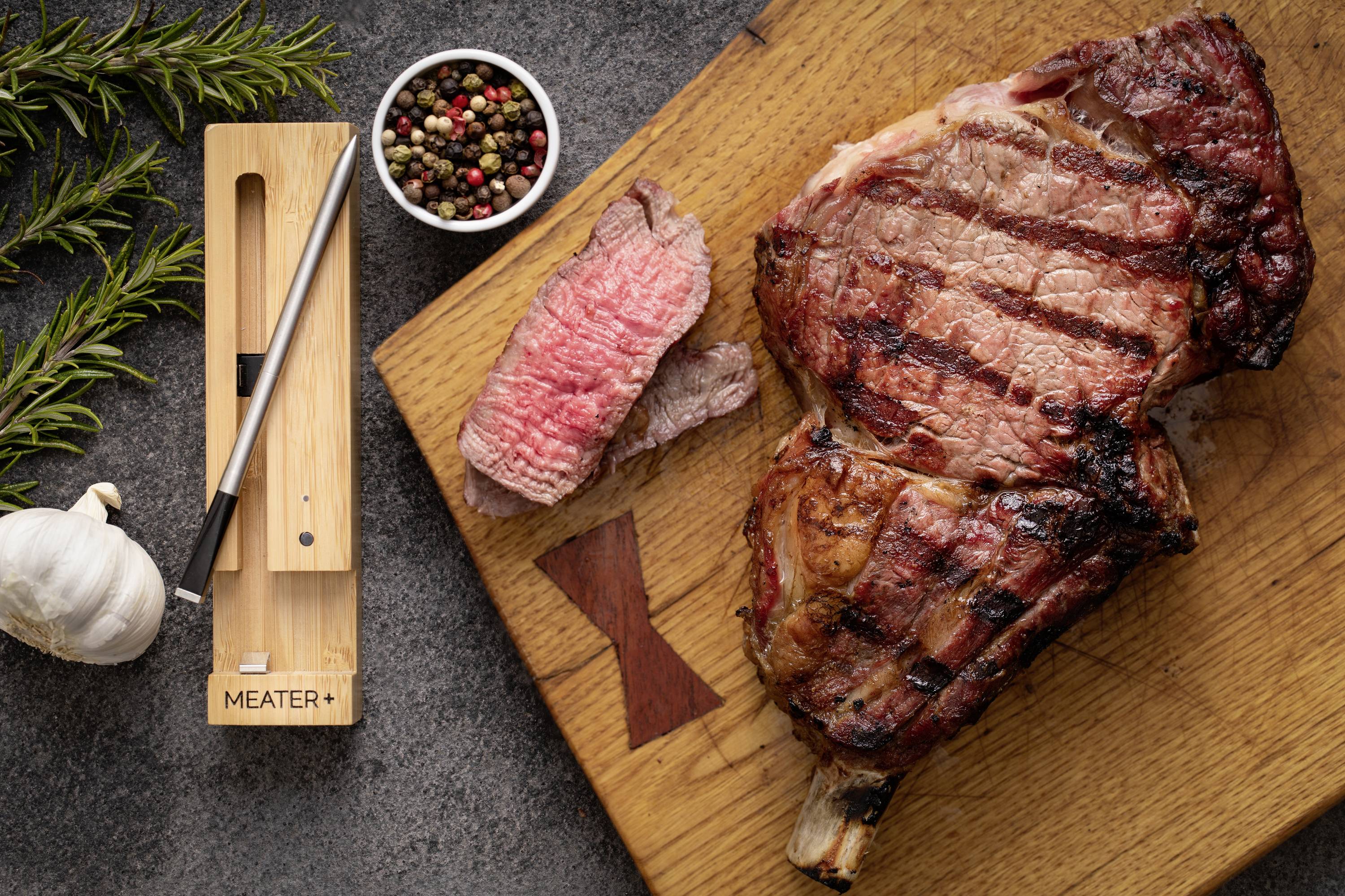 Gegrilltes Steak auf Holzbrett, daneben aufgeschnittenes Stück, Pfefferkörner, Rosmarin und Knoblauch. Meat-Thermometer links.