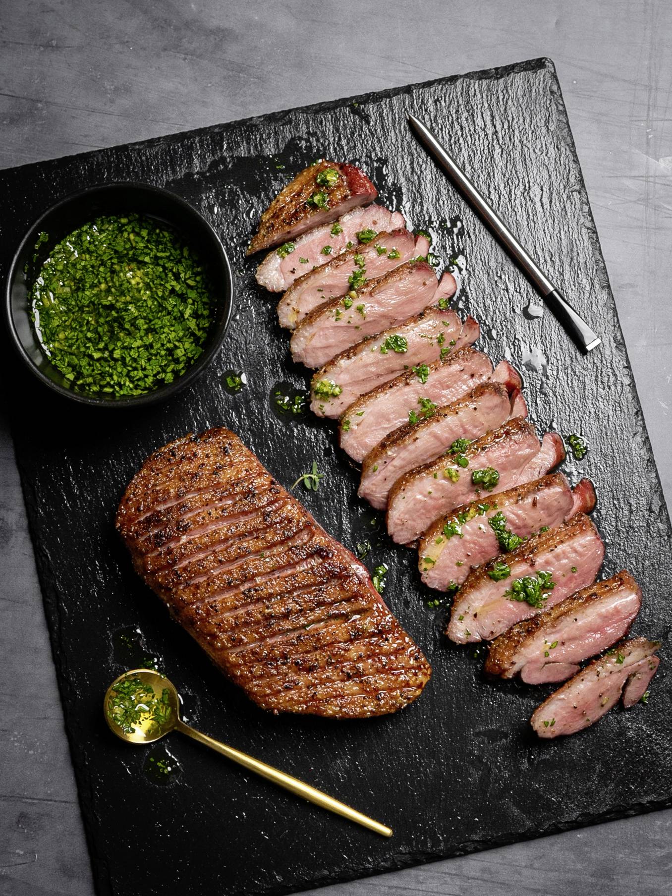 Gegrilltes Entenbrustfilet in Scheiben geschnitten, garniert mit Kräuteröl und serviert auf einer Schieferplatte mit einem kleinen Dip-Schälchen.