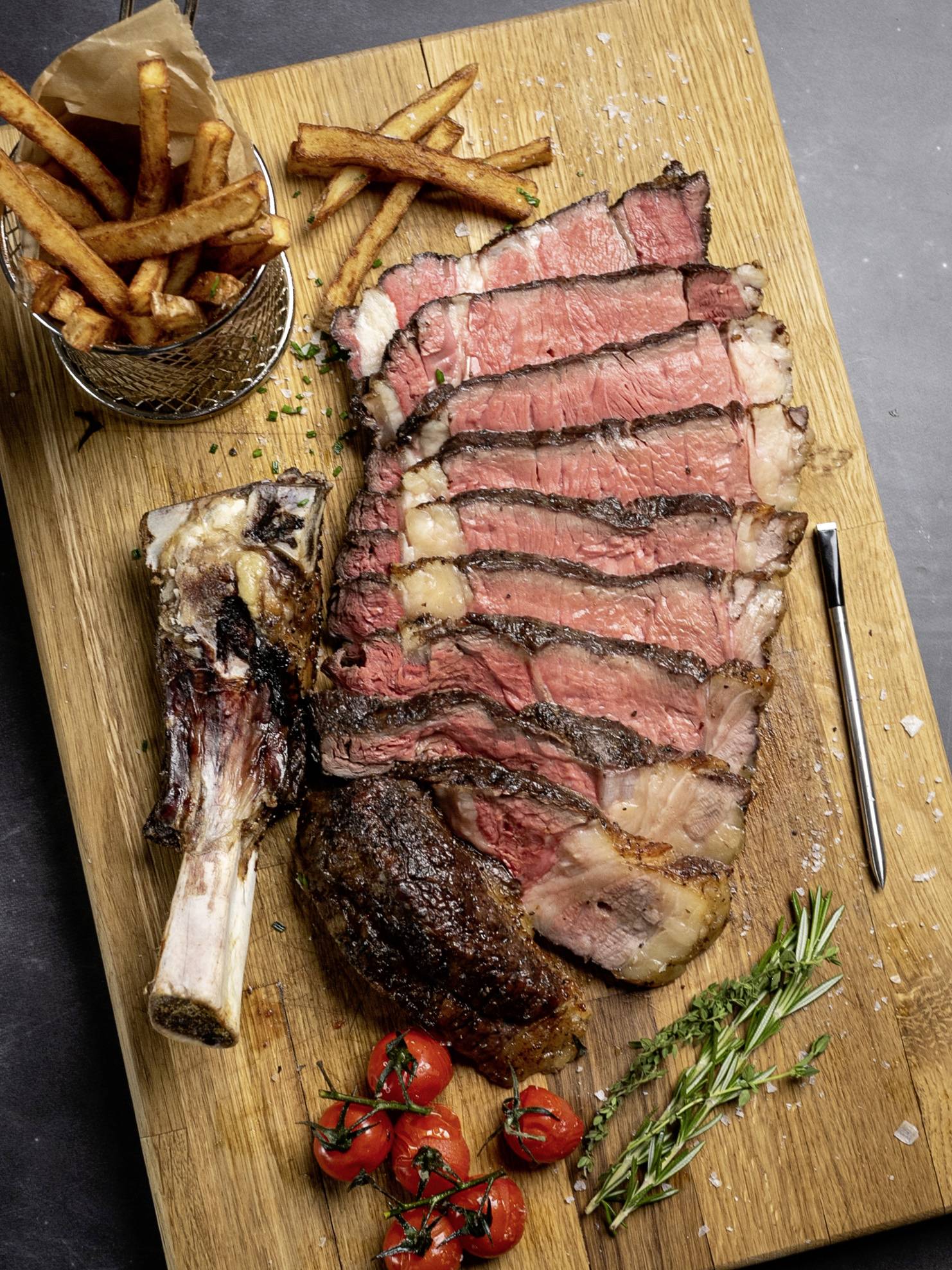 Geschnittenes Steak auf Holzplatte mit Pommes, Tomaten und Rosmarin.