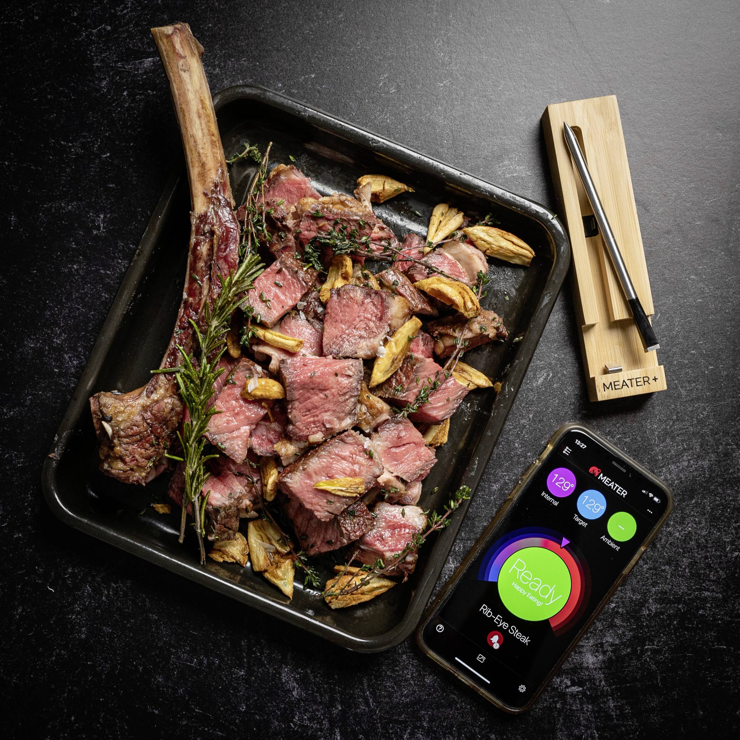 Gegrilltes Tomahawk-Steak auf einem Tablett mit Knoblauch und Rosmarin, daneben ein Thermometer und ein Handy mit Temperaturanzeige.