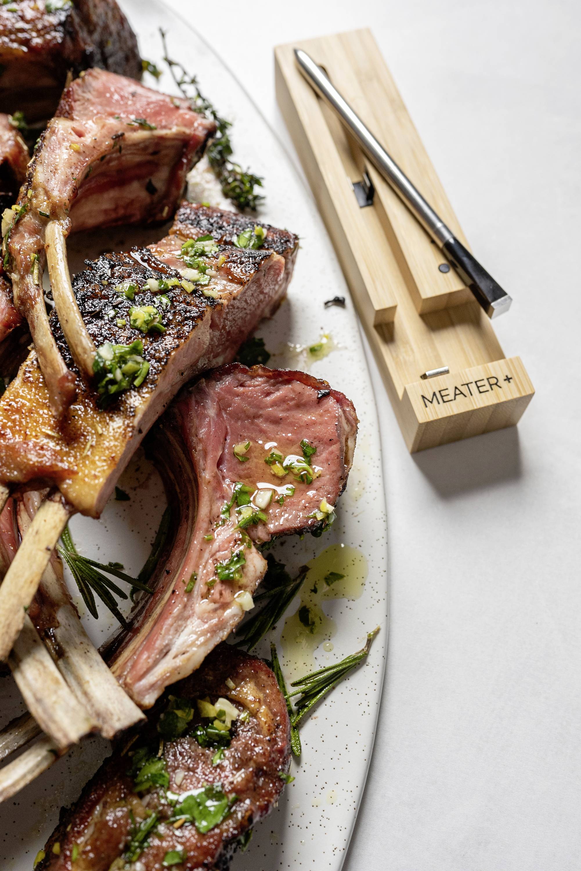 Lammkoteletts serviert auf einem Teller mit frischen Kräutern, daneben ein Fleischthermometer auf einem Holzblock.
