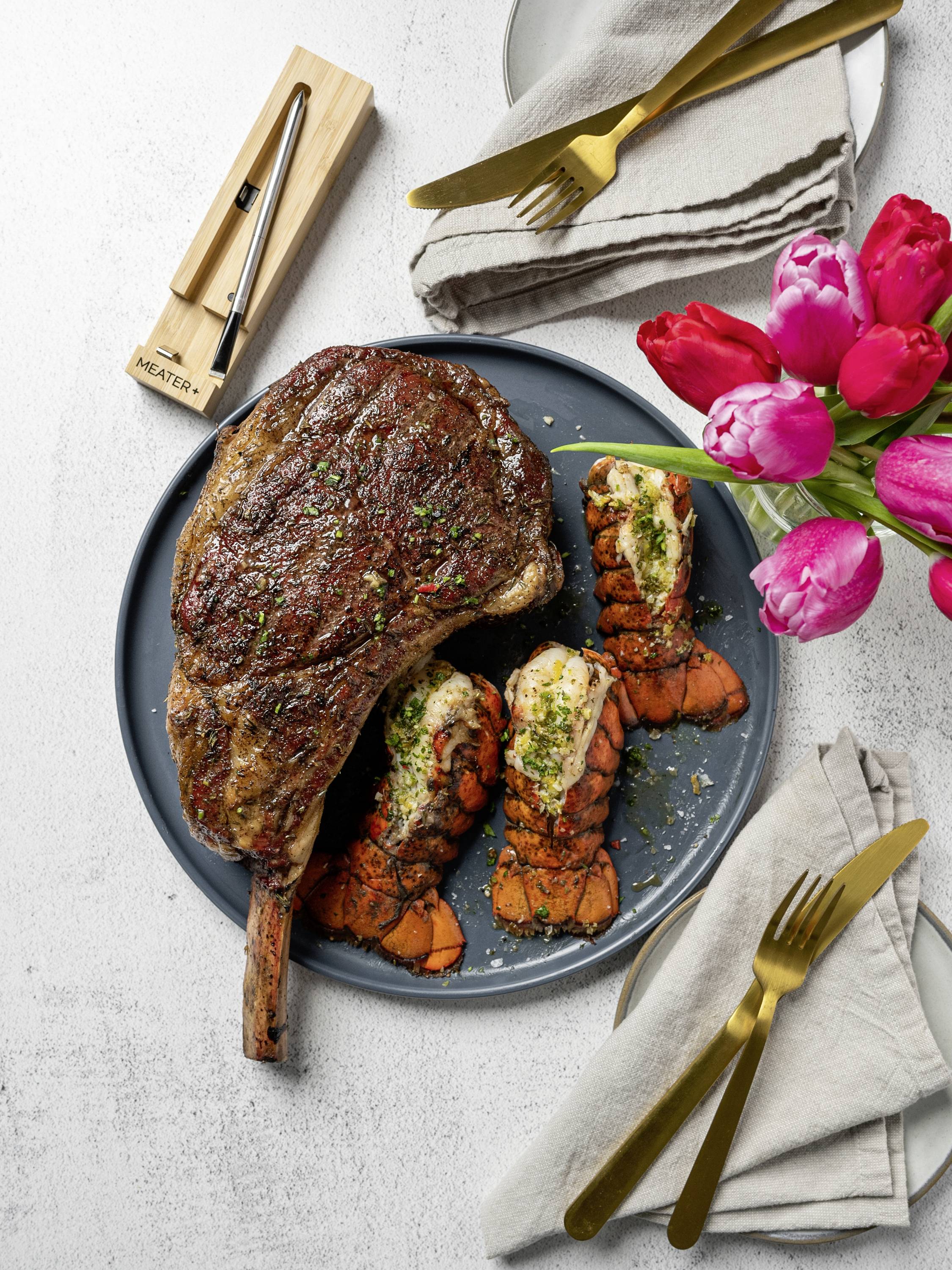 Ein Teller mit gegrilltem Steak und Hummerschwänzen, garniert mit Kräutern, daneben ein Besteckset und ein Blumenstrauß aus rosa und roten Tulpen.