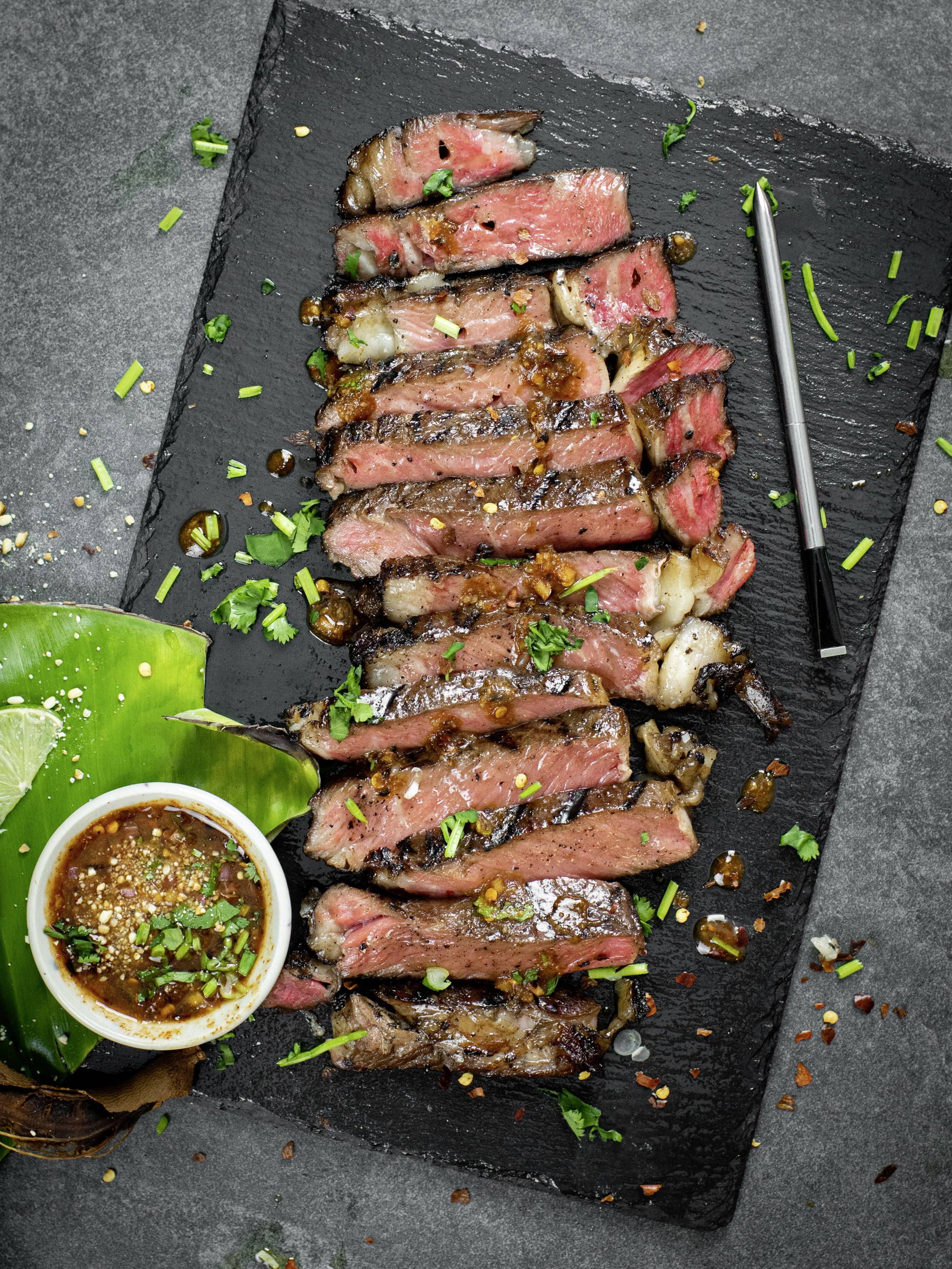 Gegrilltes Steak, in Scheiben geschnitten, serviert auf einer schwarzen Schieferplatte mit einem Becher Sauce und frischen Kräutern als Garnitur.