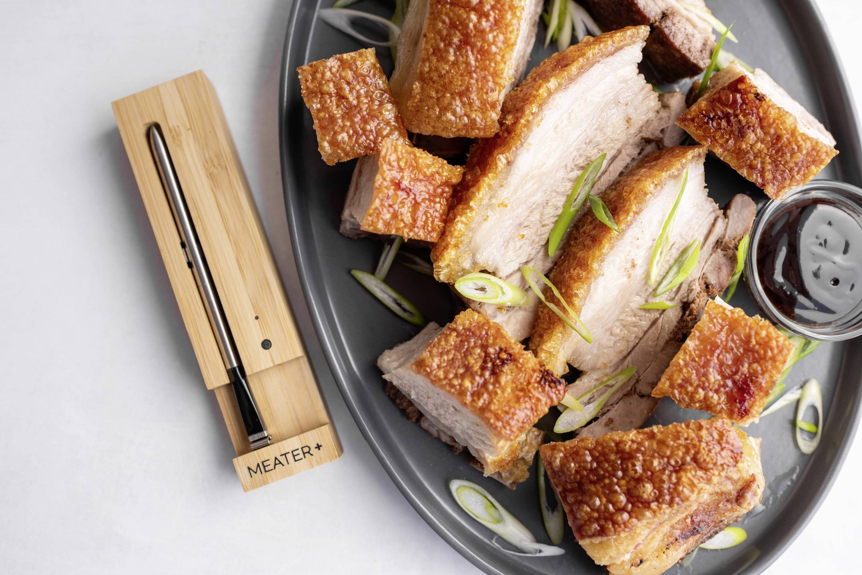 Schweinebraten mit knuspriger Kruste auf Teller, verziert mit Frühlingszwiebeln. Daneben ein digitaler Bratenthermometer.