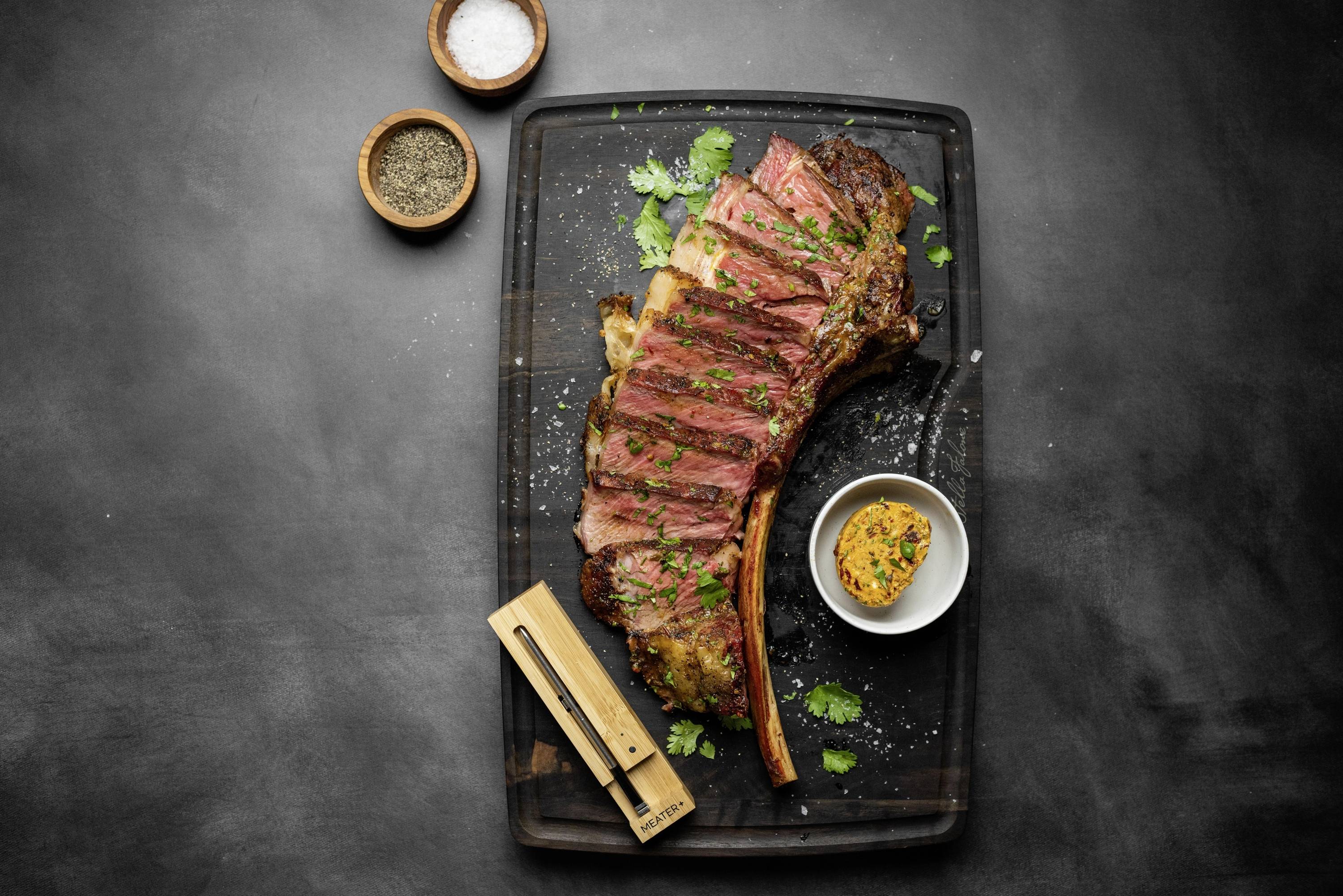 Ein gegrilltes Tomahawk-Steak auf einem Brett, in Scheiben geschnitten, mit Kräutern garniert, daneben Salz, Pfeffer und eine Gewürzbutter.