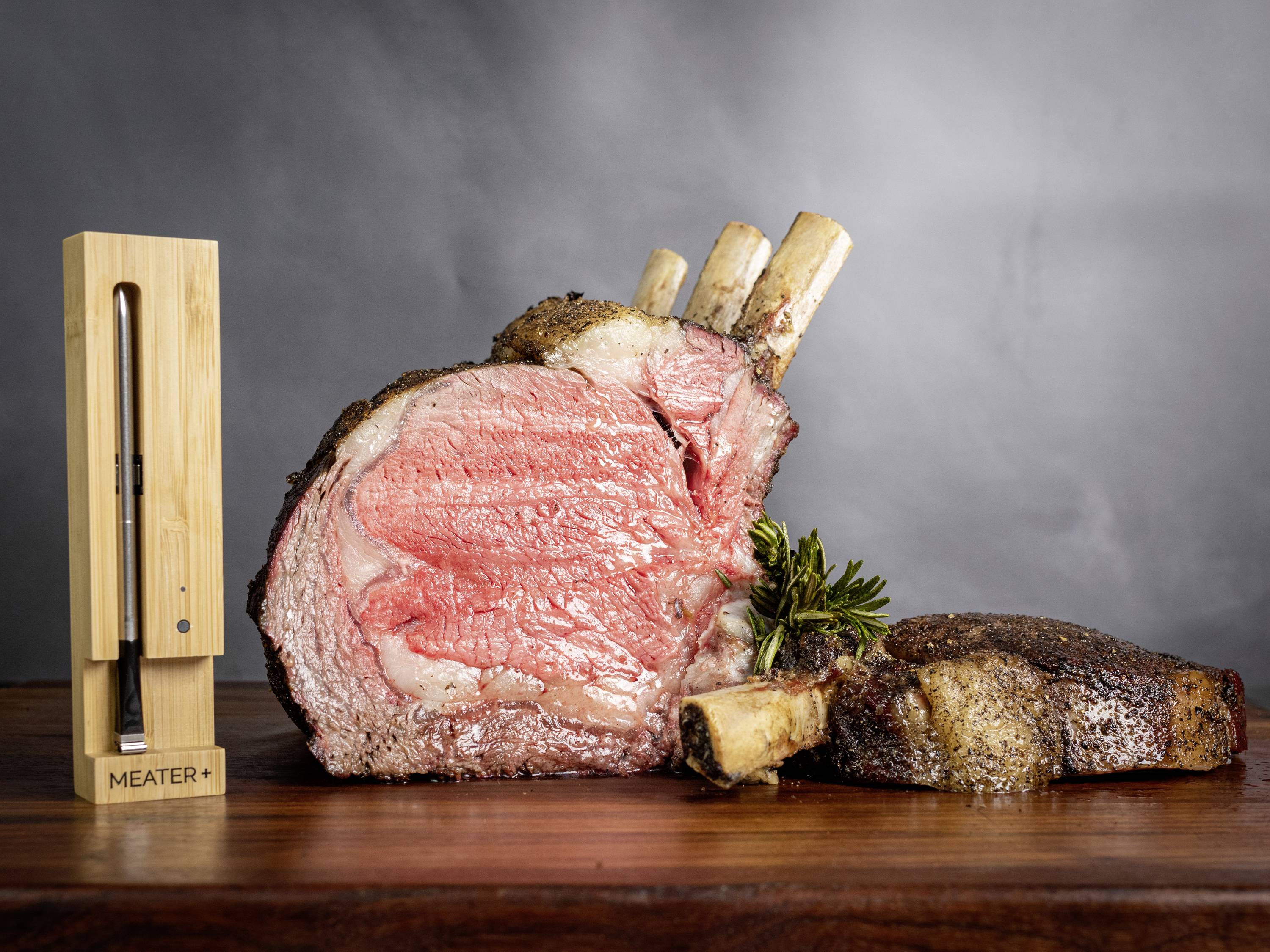 Ein großes, saftiges Stück Rindfleisch mit Knochen liegt auf einem Schneidebrett. Daneben steht ein Fleischthermometer in einem Holzständer.