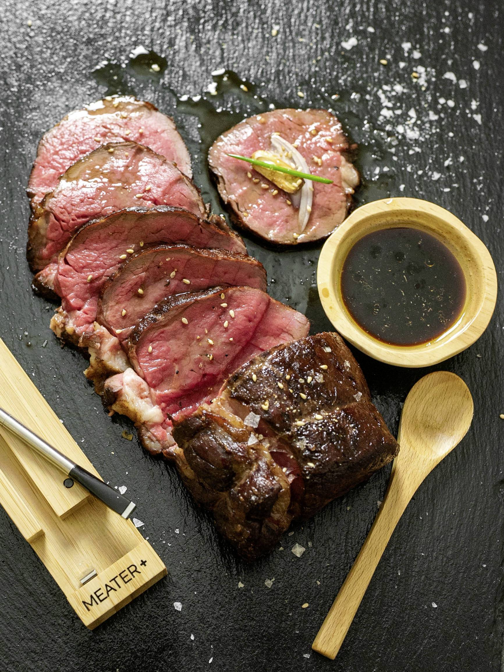 Roastbeef, in Scheiben geschnitten, auf schwarzem Schieferbrett, mit Sauce in einer Schale und einem Holzlöffel daneben.