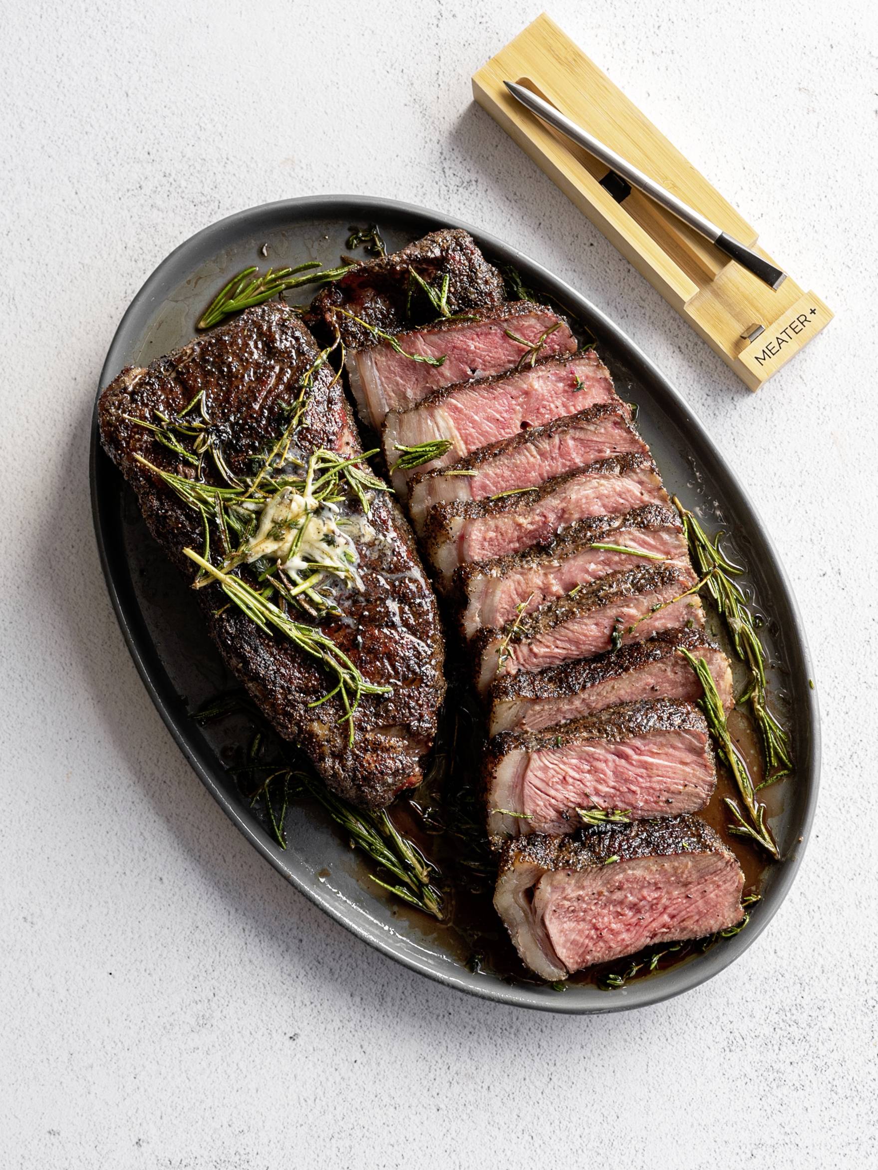 Auf einem schwarzen ovalen Teller liegen zwei saftig gebratene Steakstücke, eins ganz und eins aufgeschnitten, garniert mit Rosmarin.