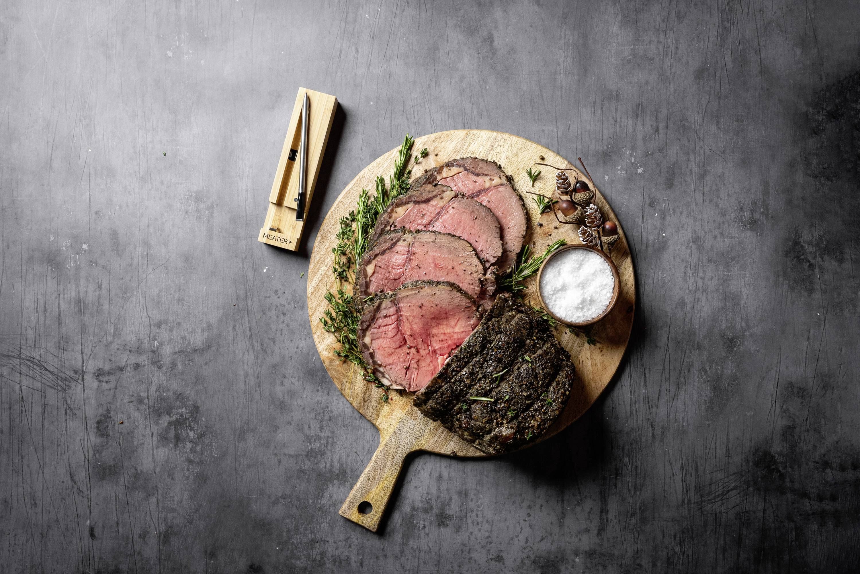 Roastbeef in Scheiben auf einem Holzbrett, mit Kräutern garniert, daneben Salz und ein Messer. Ideal für ein elegantes Abendessen.