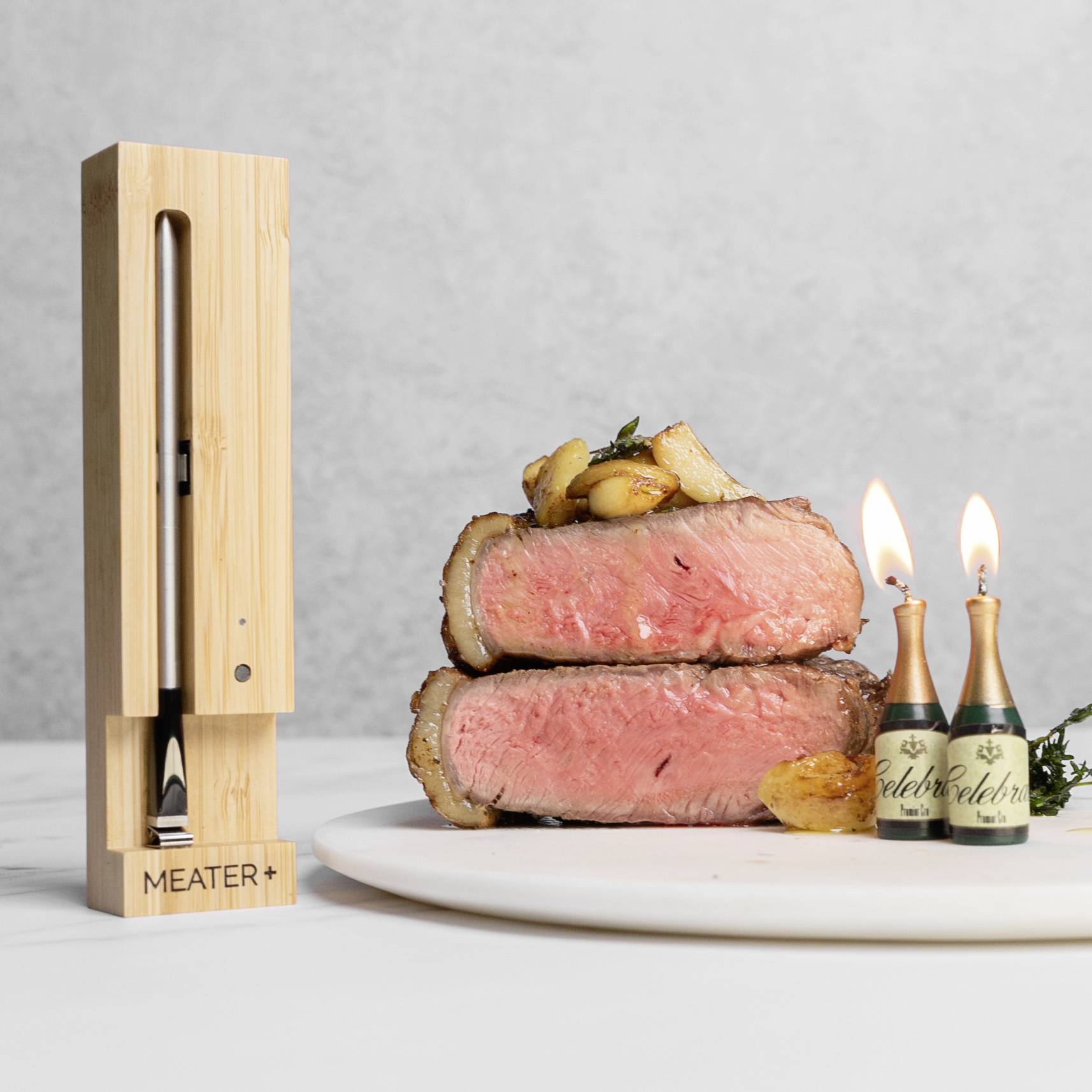 Ein Stapel medium-rare gebratenes Fleisch auf einem Teller. Daneben zwei brennende Kerzen. Links ein Holzthermometer mit 'MEATER+' Schriftzug.