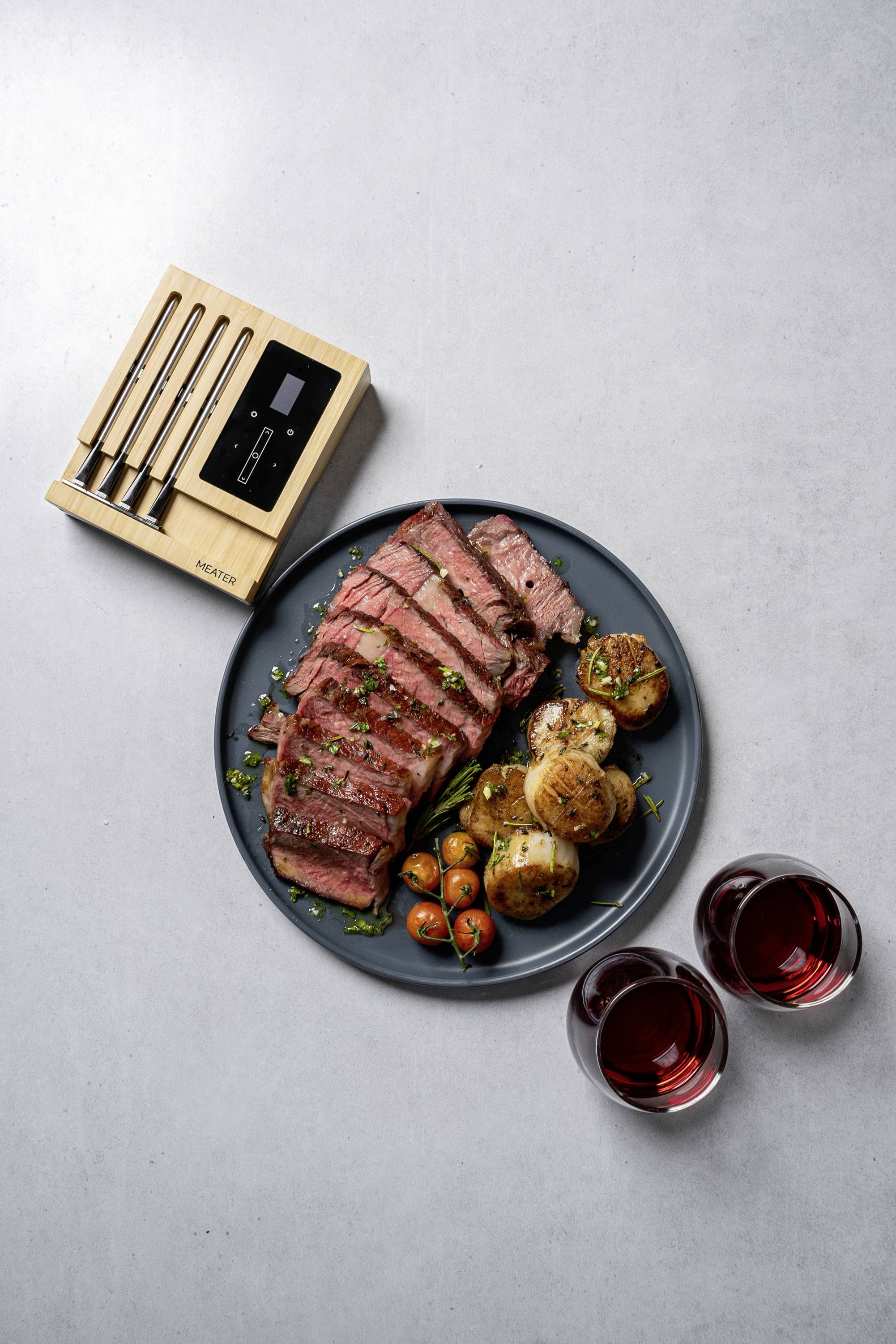Ein Teller mit geschnittenem Steak, kleinen Tomaten und Kartoffeln, flankiert von zwei Gläsern Rotwein. Ein Thermometer liegt daneben.