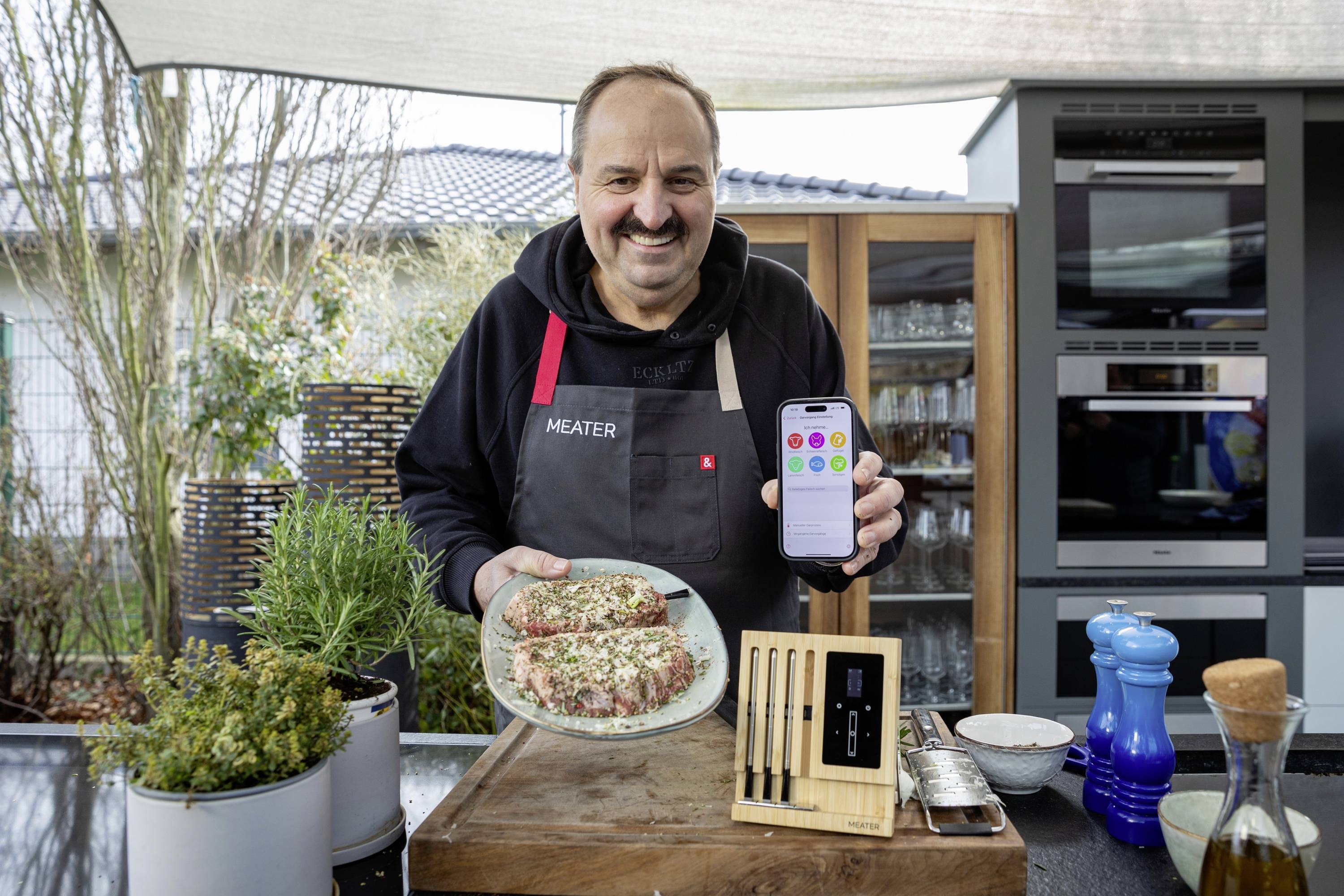 Mann im Garten mit Schürze hält Smartphone in der Hand und präsentiert gewürzte Fleischstücke auf Teller, daneben Grillthermometer.