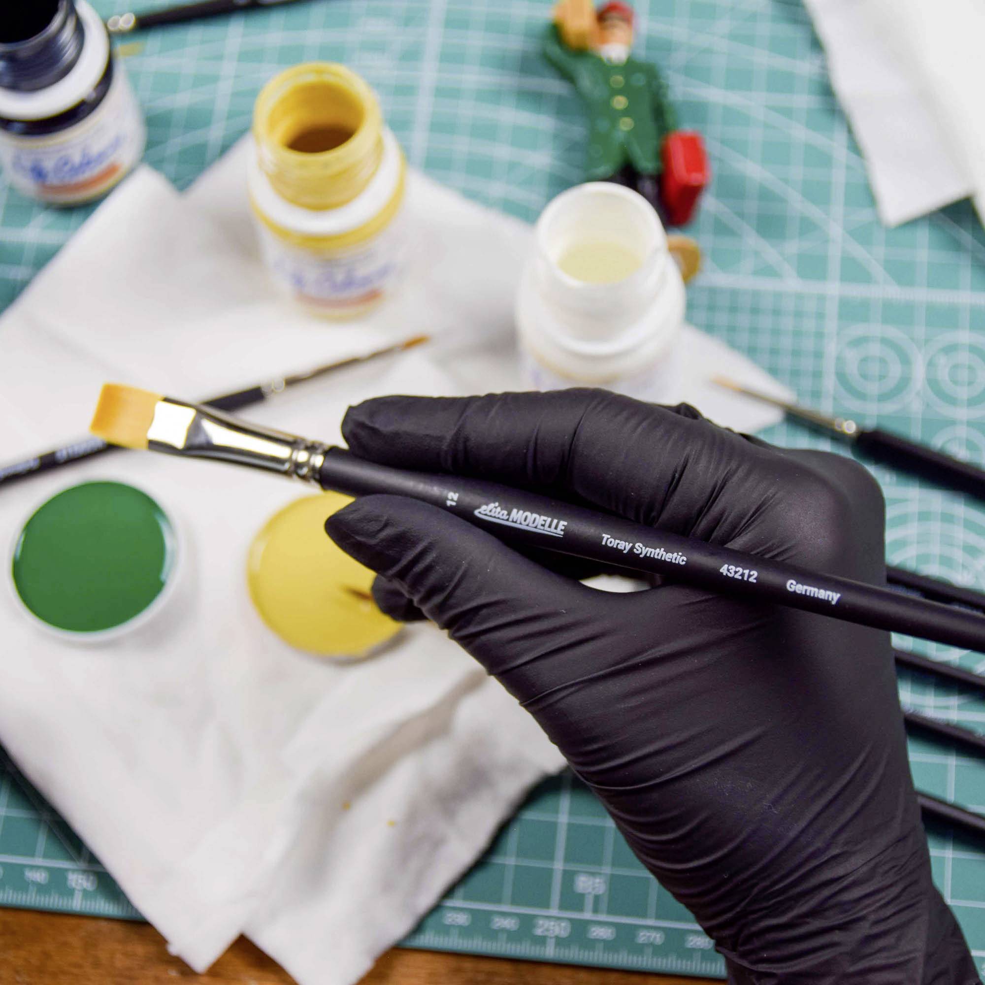 Eine Hand in Handschuhen hält einen Pinsel über einem Tisch mit bunten Farben, einem Lineal und einer kleinen Figur im Hintergrund.