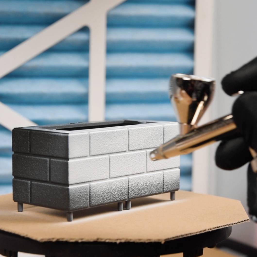 Eine Hand hält eine Airbrush und sprüht Farbe auf ein kleines Mauerstück, das auf einem Tisch steht. Hintergrund zeigt ein blaues Gittermuster.
