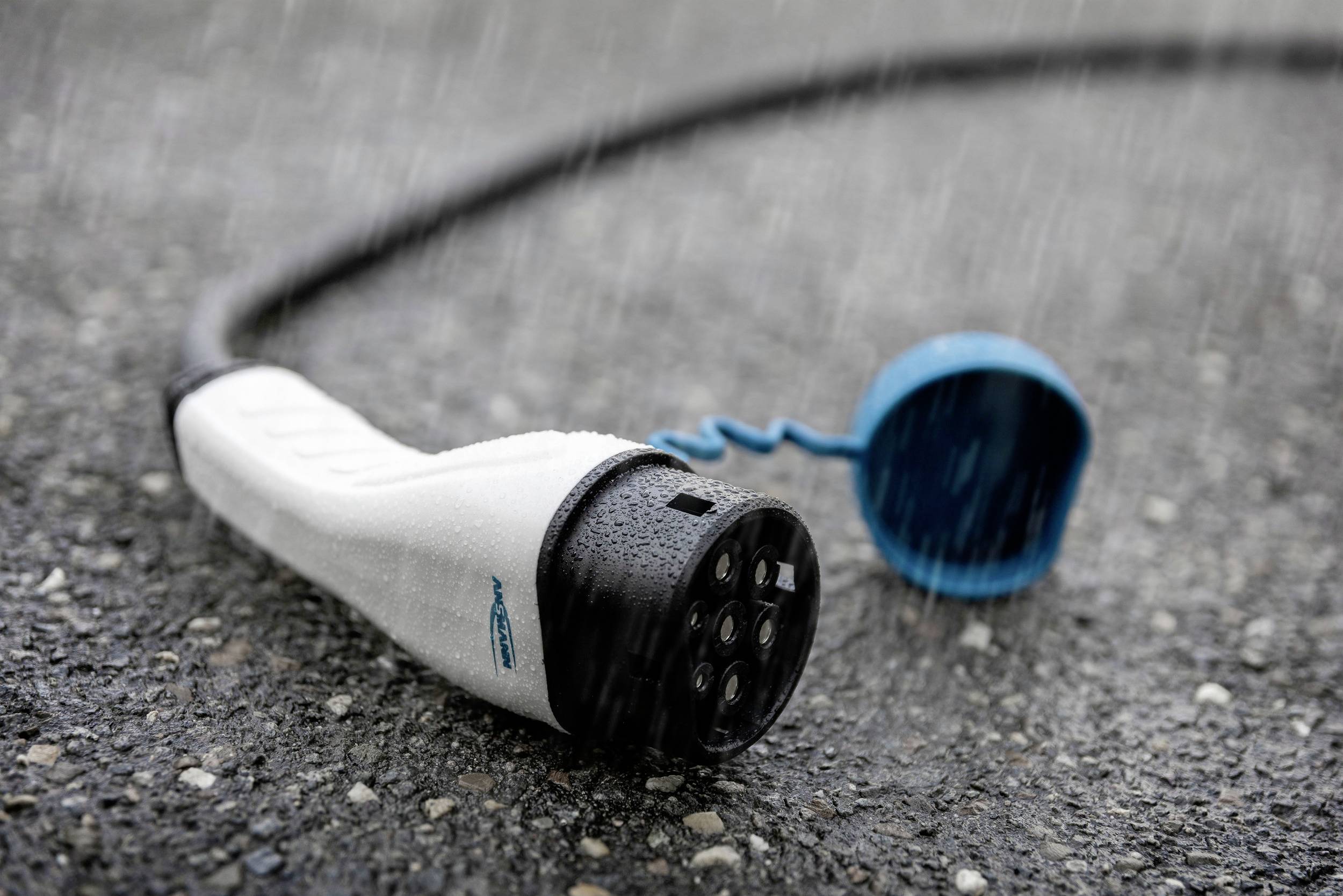 Ein Elektroauto-Ladestecker liegt im Regen auf nassem Asphalt, dessen Steckkontakte geschützt und unbenutzt sind.