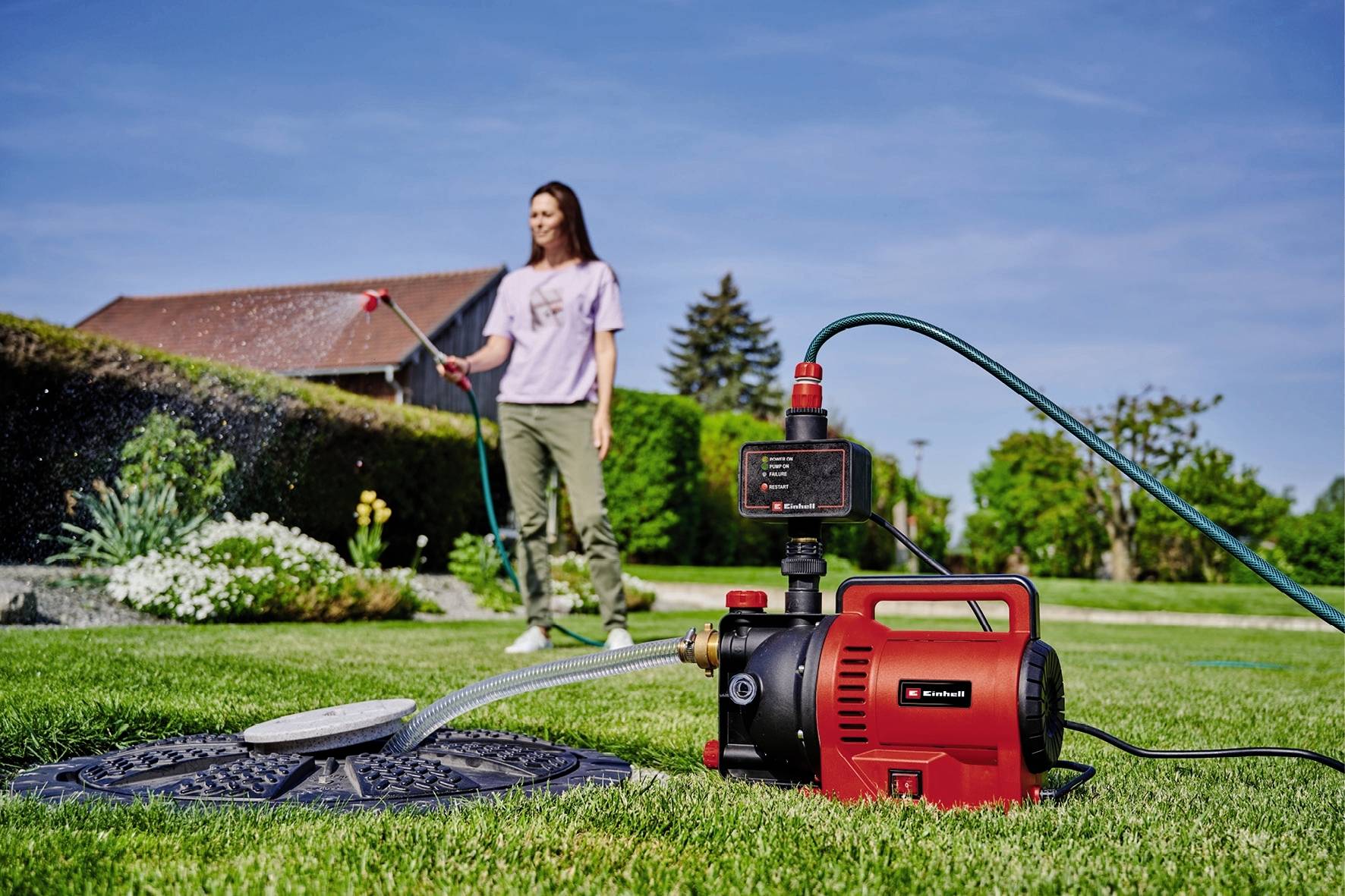 Eine Frau bewässert den Rasen im Garten mit einem roten Wasserpumpsystem im Vordergrund. Häuser und Pflanzen sind im Hintergrund sichtbar.