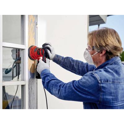 Ein Mann schleift den Holzrahmen eines Fensters mit einer elektrischen Schleifmaschine ab. Er trägt Schutzbrille und Maske.