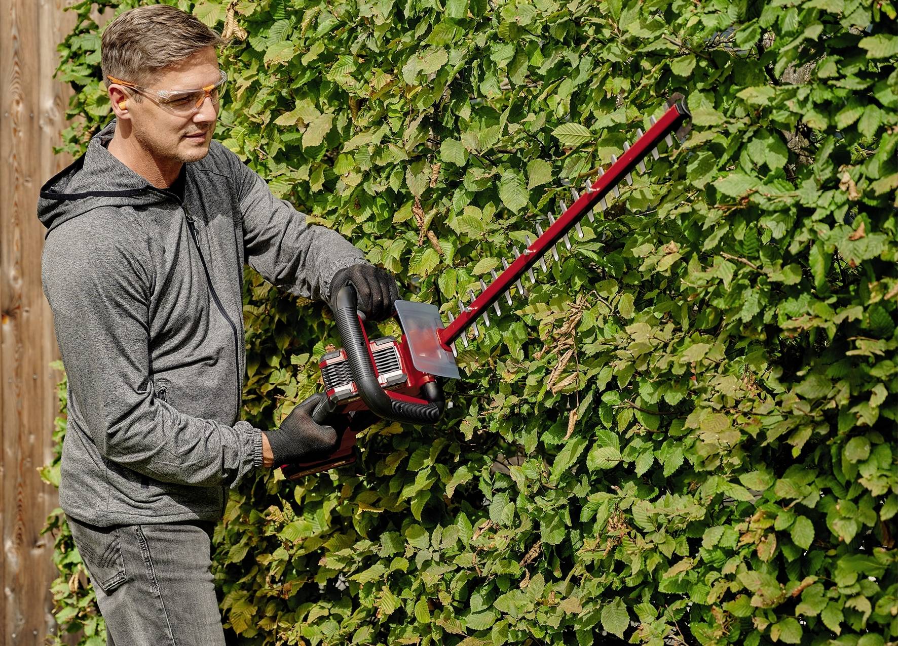 Mann schneidet mit einer elektrischen Heckenschere eine grüne Hecke. Trägt Schutzbrille und Handschuhe.