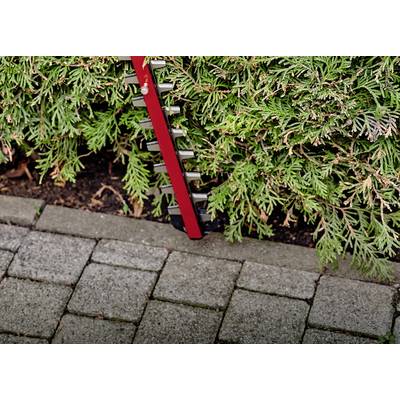 Eine rote Heckenschere schneidet eine grüne Hecke entlang eines gepflasterten Weges.