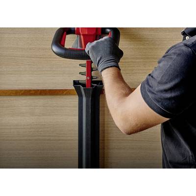 Eine Person mit schwarzem Hemd und Handschuhen bedient eine Maschine mit roten und schwarzen Komponenten vor einer Holzwand.