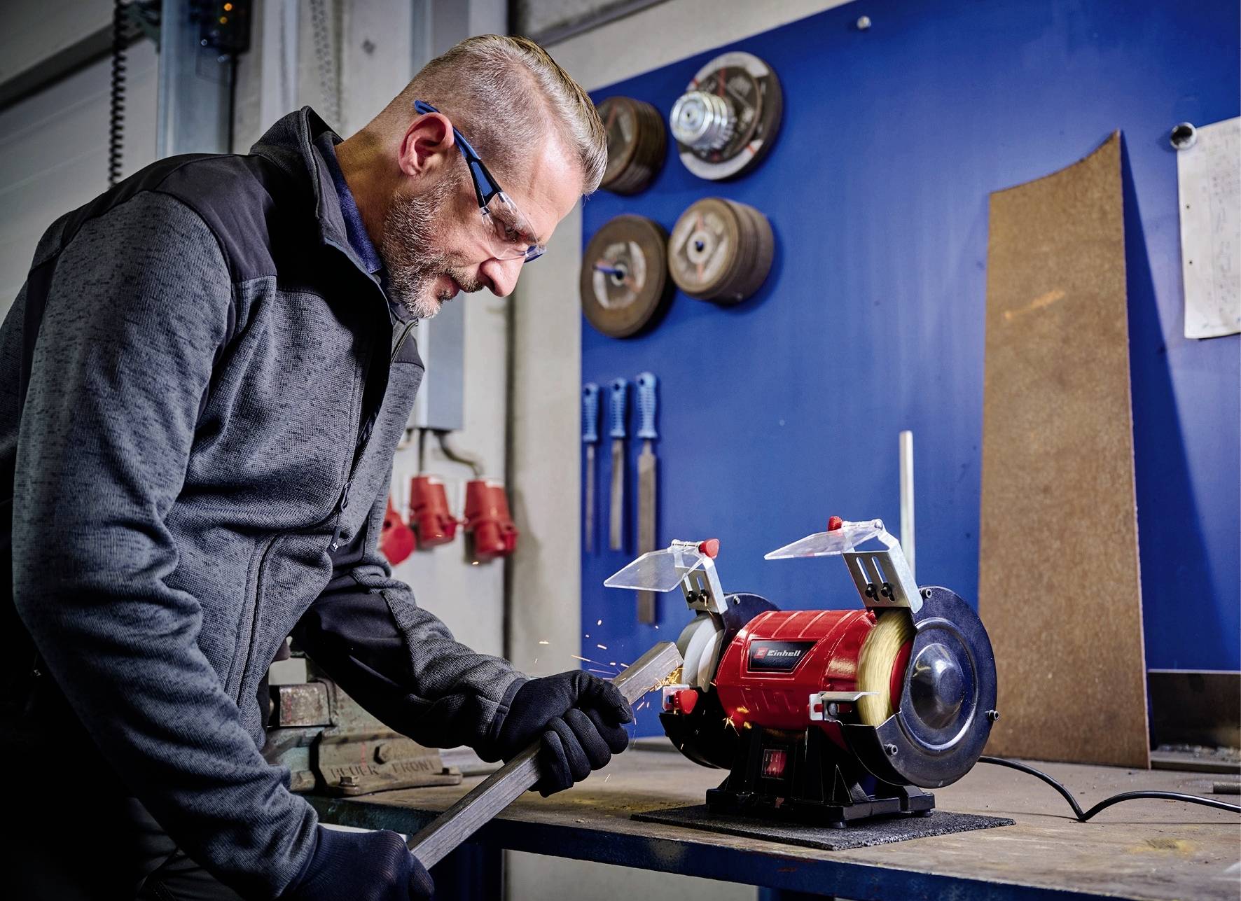Ein Mann bearbeitet ein Metallstück mit einer Schleifmaschine in einer Werkstatt. Funken fliegen, Schutzbrille und Handschuhe sind sichtbar.
