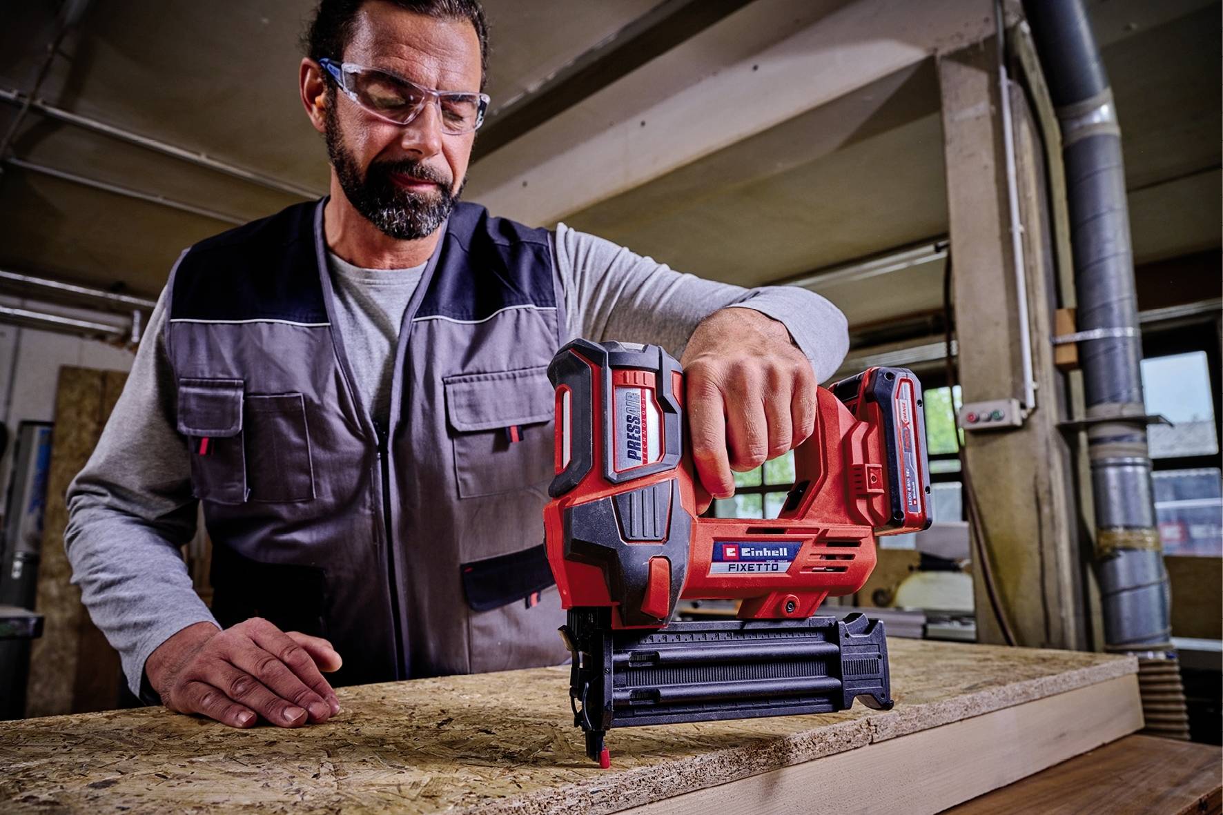 Ein Mann in Schutzbrille und Arbeitskleidung benutzt ein Nagelgerät, um Holzplatten in einer Werkstatt zu befestigen.
