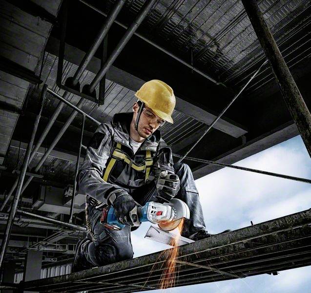 Ein Arbeiter mit Schutzhelm und Sicherungsgurt schneidet Metall mit einem Winkelschleifer auf einer Baustelle. Funken fliegen.