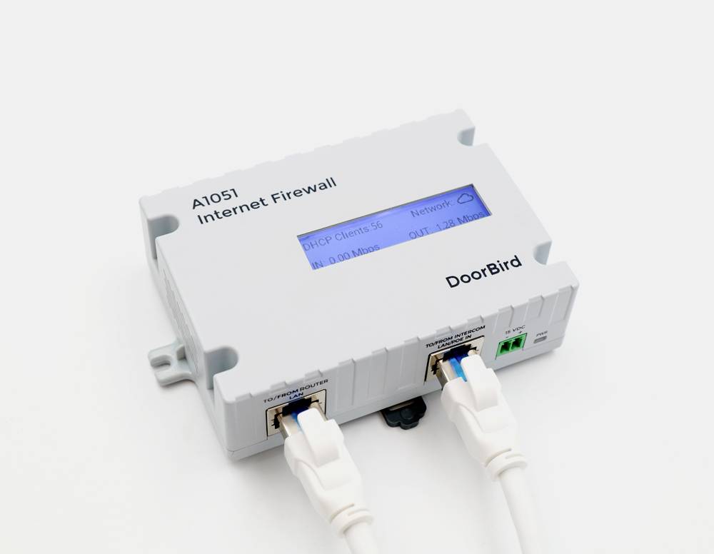 Ein weißes Gerät mit der Aufschrift 'A1051 Internet Firewall' und 'DoorBird'. Es hat zwei angeschlossene Kabel und ein LCD-Display mit Netzwerkhinweisen.