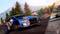 Ein blaues Rallyeauto fährt schnell auf einer kurvigen Schotterstraße. Im Hintergrund sind Bäume und ein Berg zu sehen, rechts steht das Logo von 'V-Rally 4'.