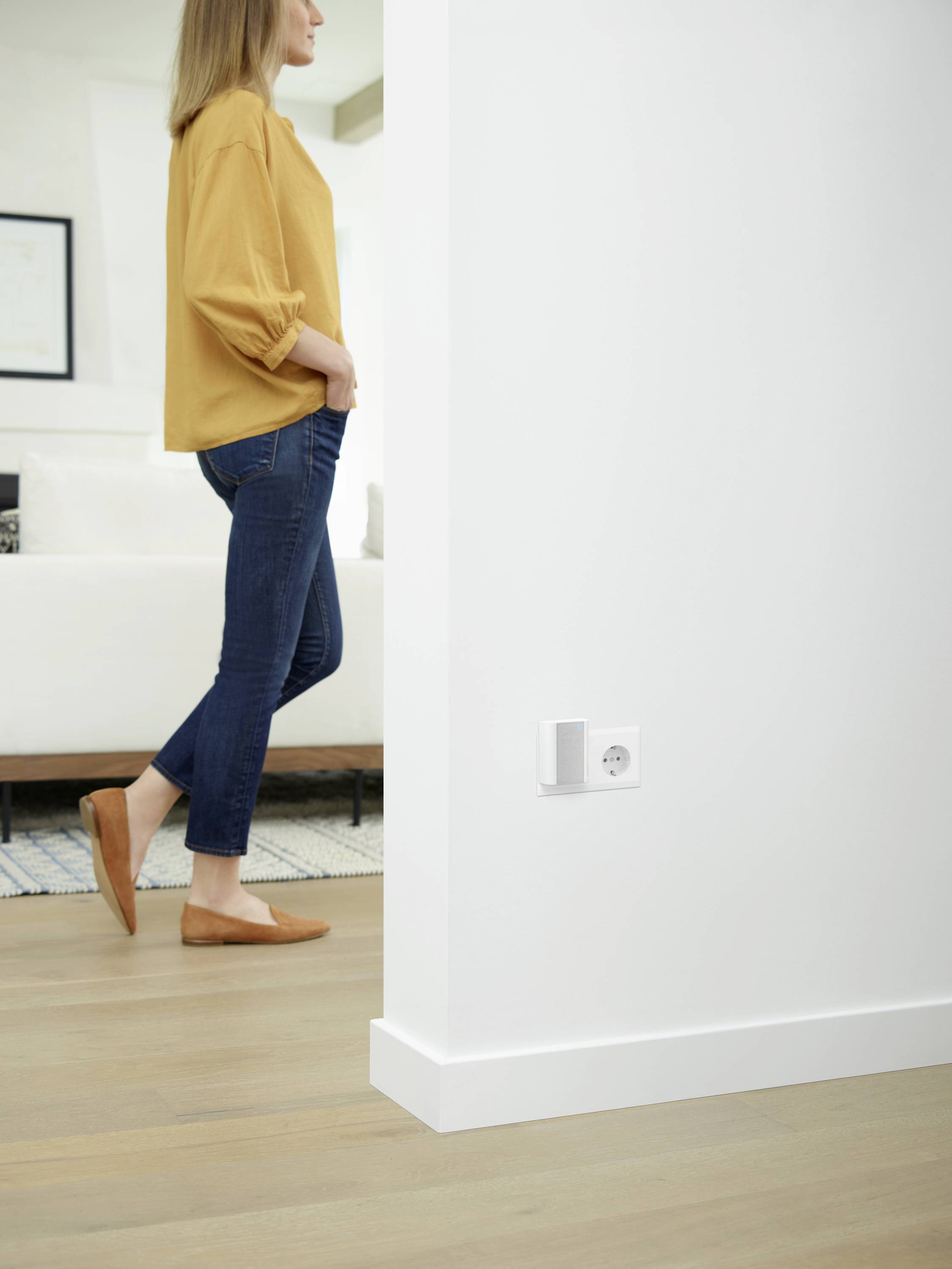 Eine Person geht durch ein modernes Wohnzimmer mit Holzboden. Eine Wandsteckdose und ein kleines, rechteckiges Gerät sind an der Wand angebracht.