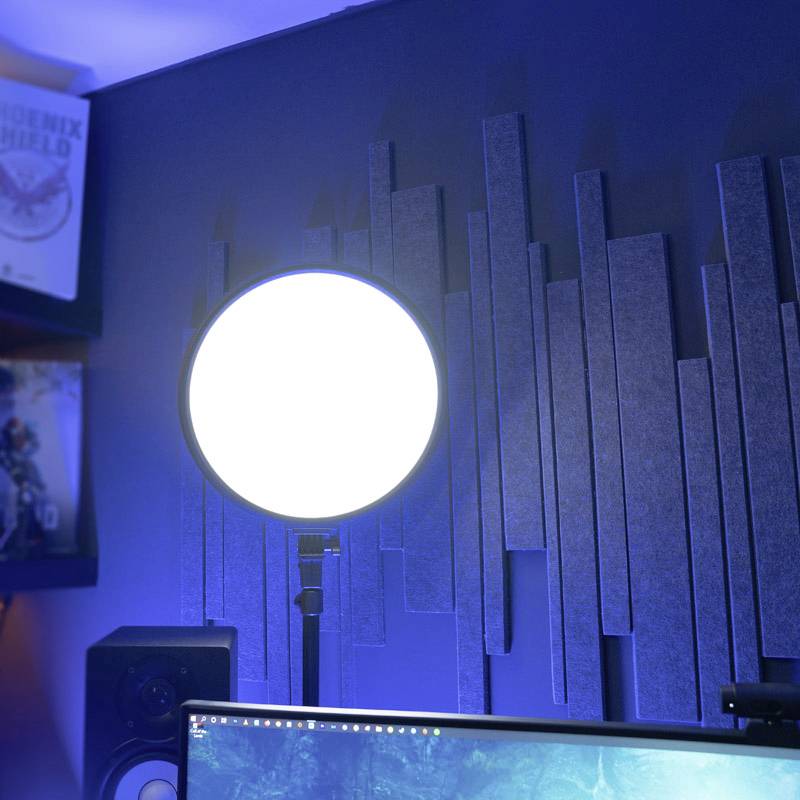 Ein runder, heller Studiolichtschirm leuchtet an einer Wand mit schallabsorbierenden Paneelen, neben einem Monitor und Lautsprechern.