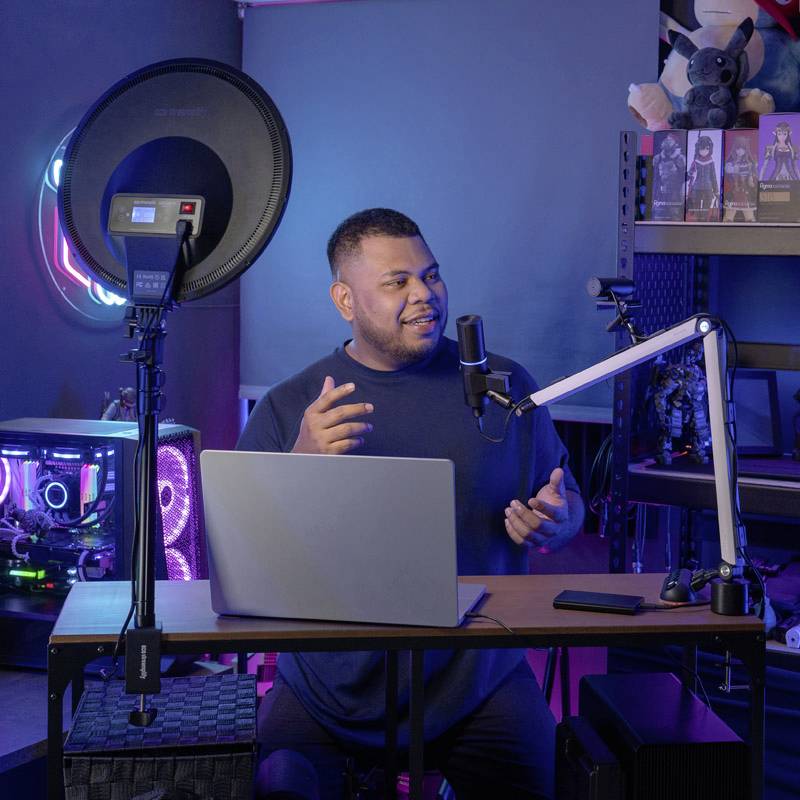 Eine Person hält einen Vortrag in einem abgedunkelten Raum, vor einem Laptop mit Mikrofon, Ringlicht und Regalen mit Figuren im Hintergrund.