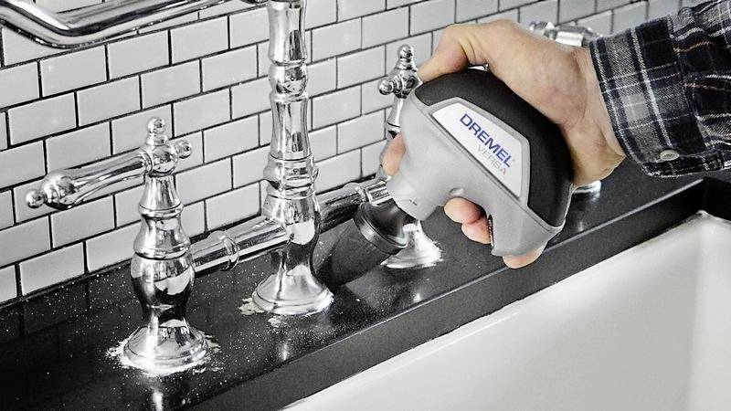 'Person verwendet ein elektrisches Reinigungswerkzeug, um Wasserhähne in einem Badezimmer mit Fliesenwand zu reinigen.'
