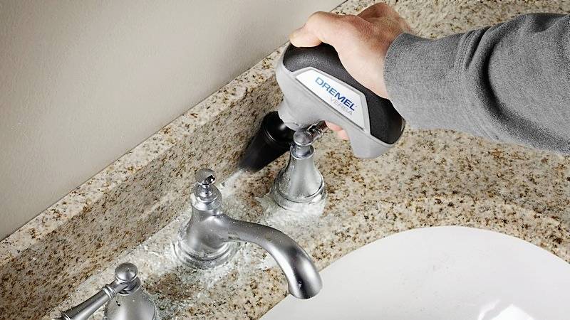 Eine Person reinigt ein Waschbecken mit einem Handpolierer auf einer Steinplatte. Der Polierer entfernt Schmutz um den Wasserhahn herum.