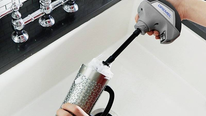 Eine Person reinigt einen silbernen Becher in einem Waschbecken mit einer elektrischen Reinigungsbürste.