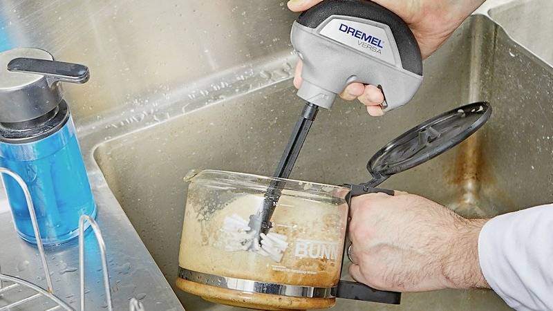 Eine Person reinigt eine verschmutzte Kaffeekanne in einem Waschbecken mit einem Dremel-Reinigungsgerät.