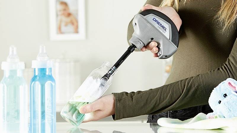 Eine Person reinigt eine Babyflasche mit einem speziellen Gerät. Im Hintergrund sind weitere Flaschen und ein Stofftier zu sehen.