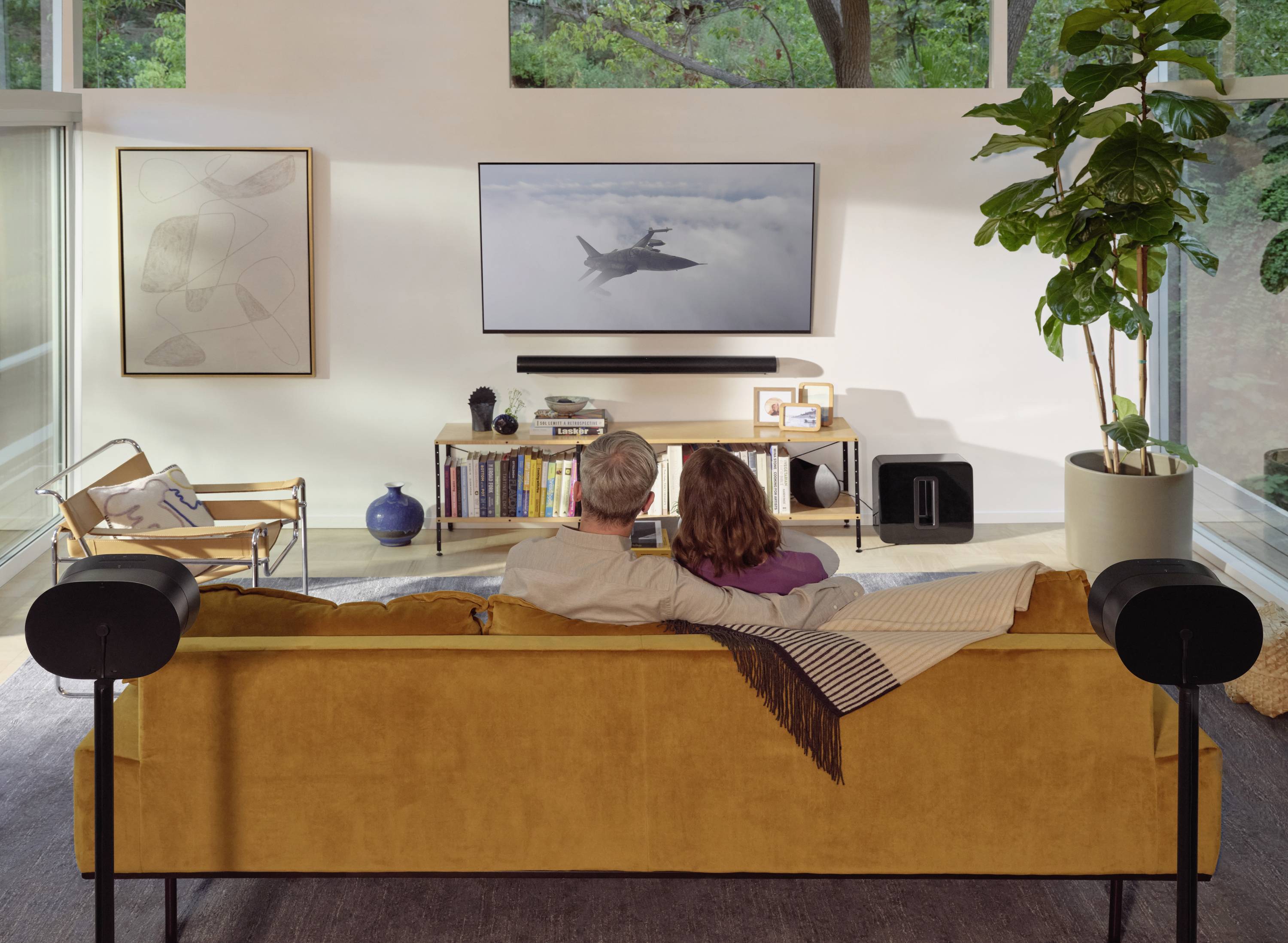Ein älteres Paar sitzt auf einem Sofa und schaut gemeinsam einen Fernseher an, auf dem ein Bild eines Flugzeugs im Flug zu sehen ist.