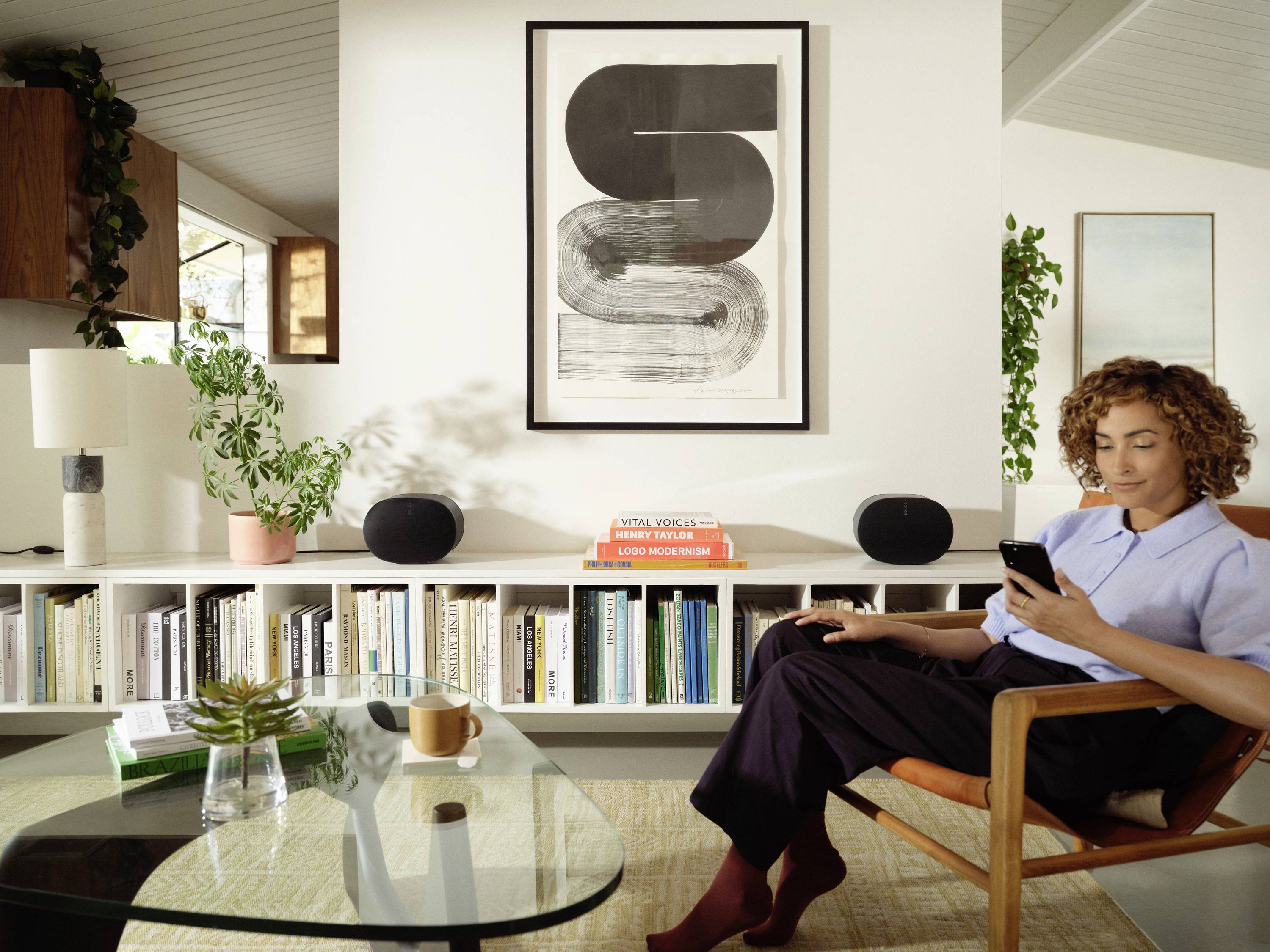 Eine Frau sitzt in einem modern eingerichteten Wohnzimmer und schaut auf ihr Handy. Im Hintergrund sind Bücherregale und ein Kunstwerk an der Wand zu sehen.