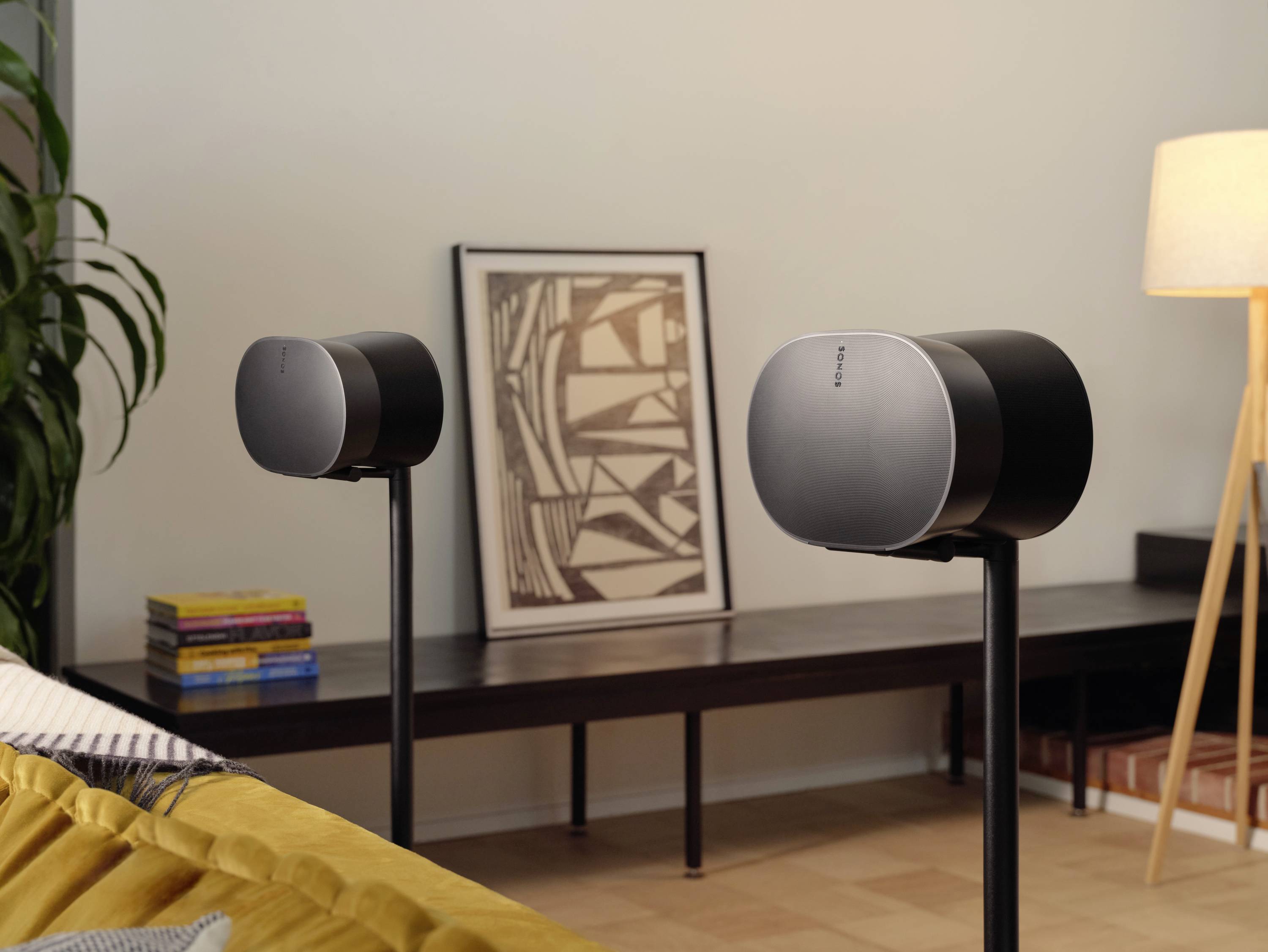 Zwei Lautsprecher stehen im Wohnzimmer auf Ständern. Im Hintergrund befinden sich ein Bild, Bücher und eine Lampe.