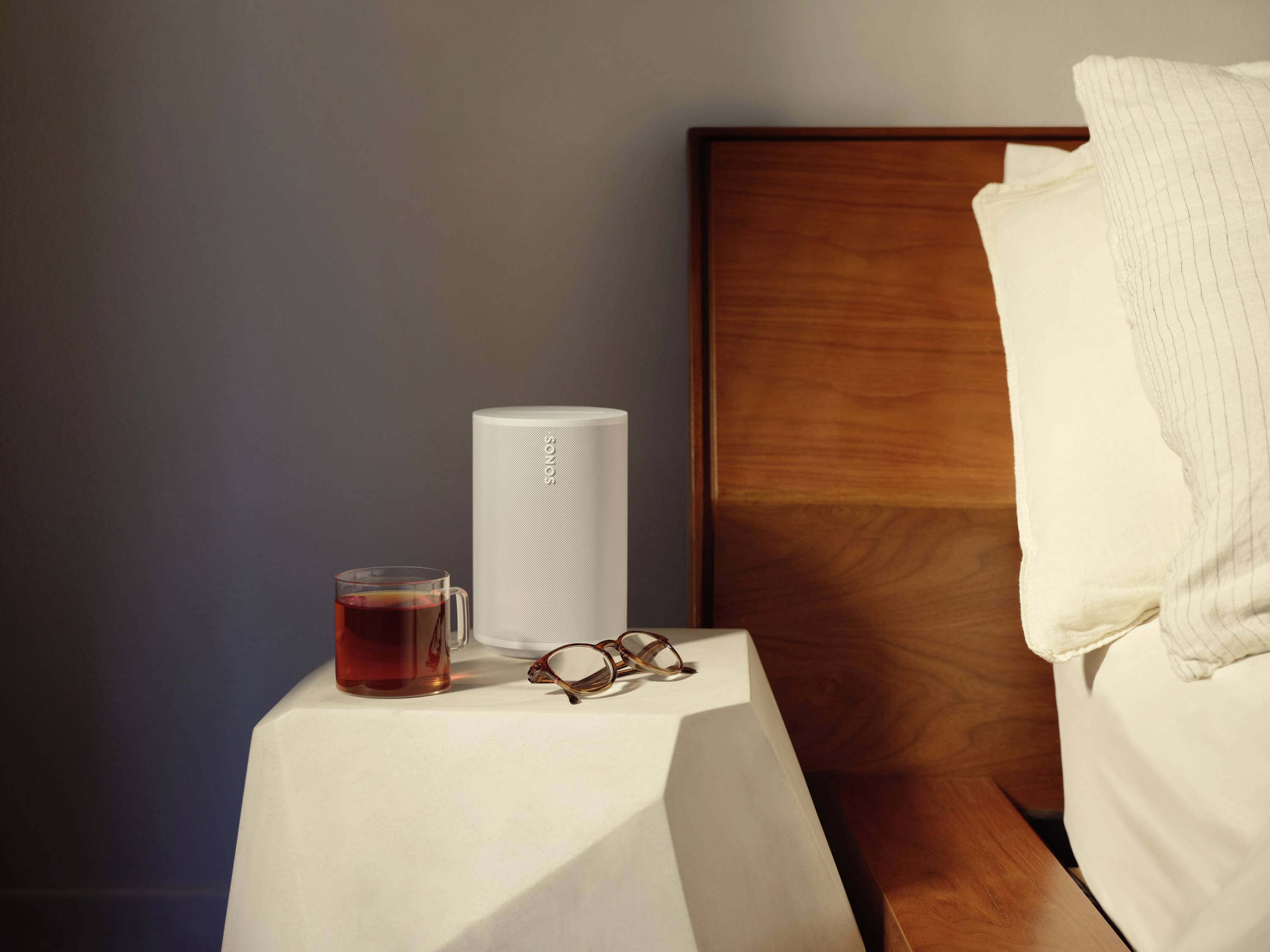 Ein weißer Lautsprecher steht auf einem Nachttisch neben einer Tasse Tee und einer Brille. Im Hintergrund ist ein Bett zu sehen.